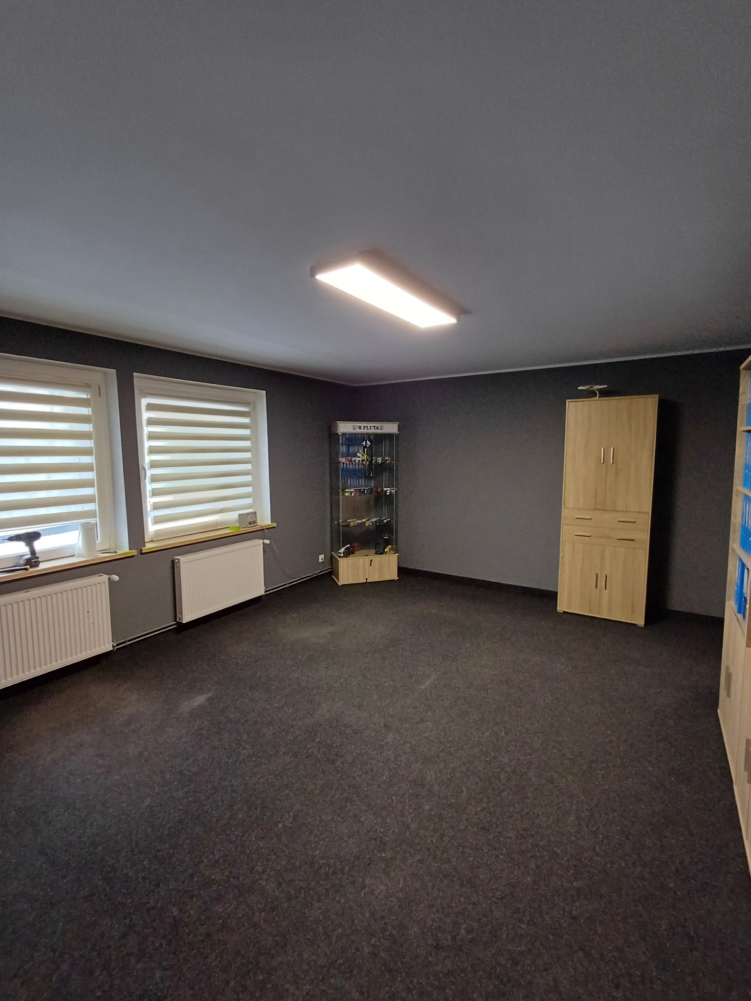 Pomalowane na szaro wnętrze pokoju z oknami z żaluzjami, grzejnikami, szafką ekspozycyjną i szafą. Na parapecie wiertarka akumulatorowa.