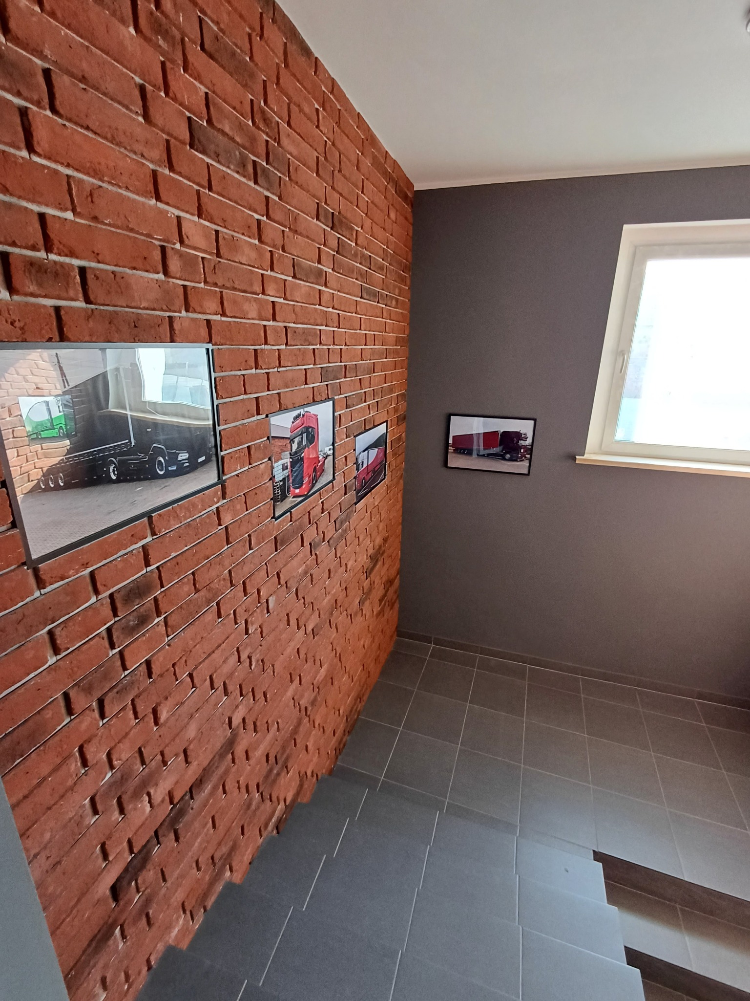Korytarz ze schodami i ceglaną ścianą, na której wiszą oprawione zdjęcia ciężarówek. Szare ściany i podłoga, okno z białymi ramami.