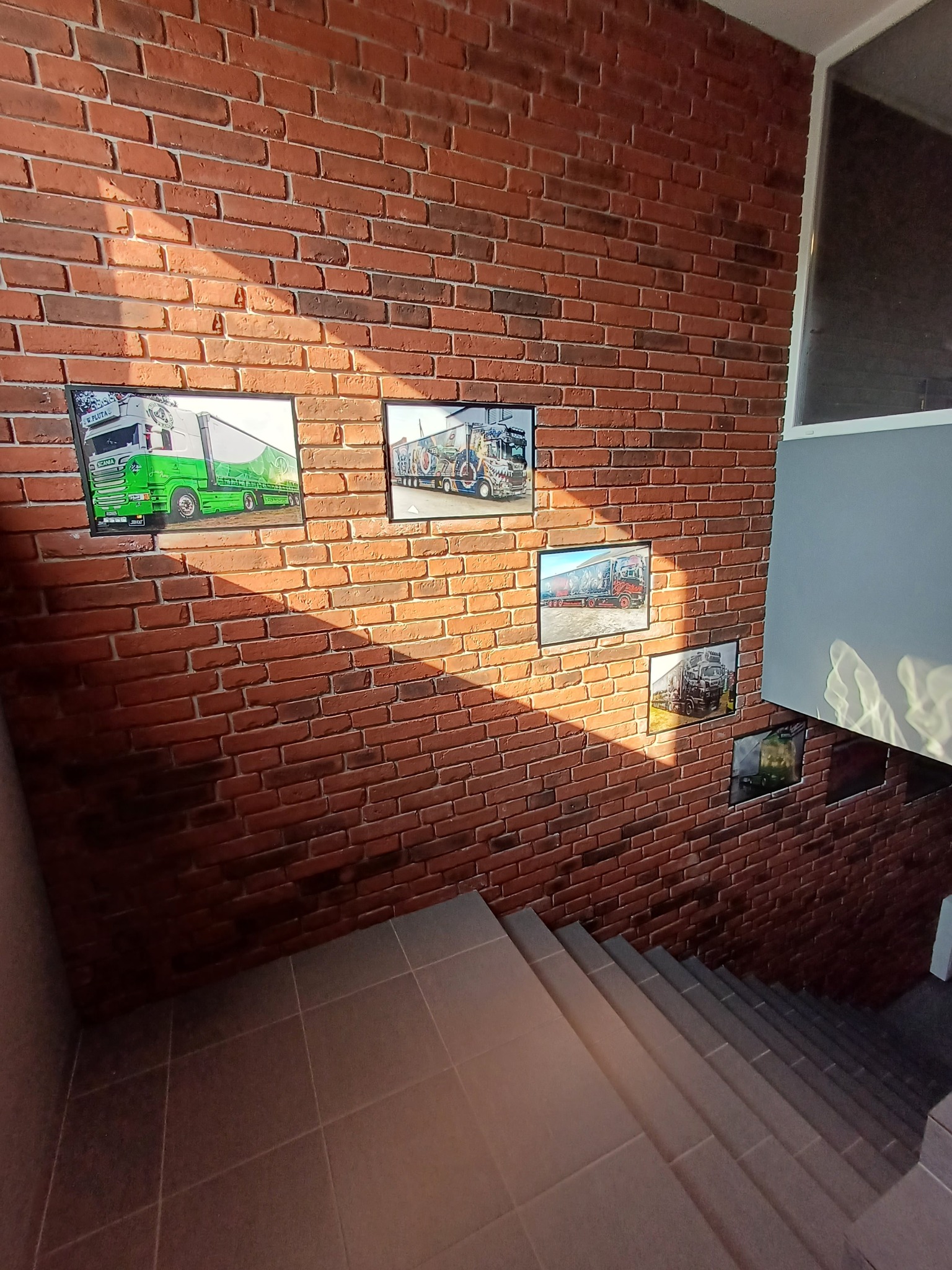 Ceglana ściana z dekoracyjnymi obrazami ciężarówek, oświetlona promieniami słońca, widok ze schodów. Nietypowe wykończenie wnętrza z motywem transportowym.