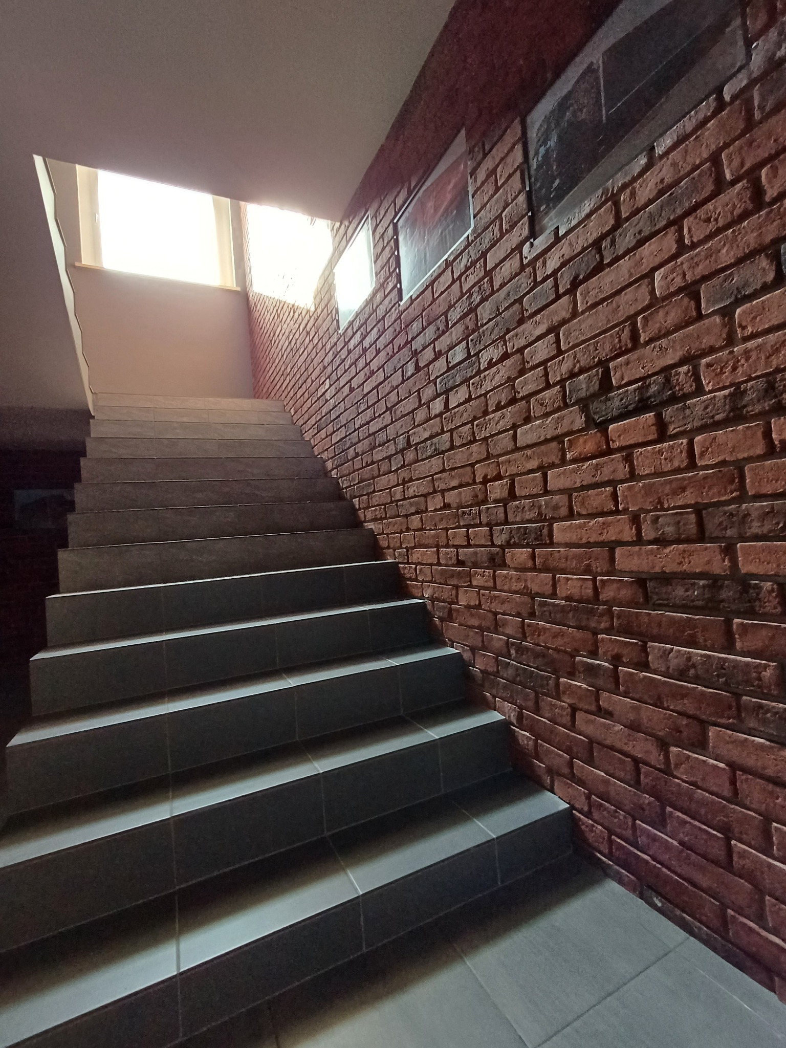 Klatka schodowa z szarymi płytkami na stopniach i ścianą z czerwonej cegły. Na ścianie wiszą obrazy. W górze okno wpuszcza naturalne światło.