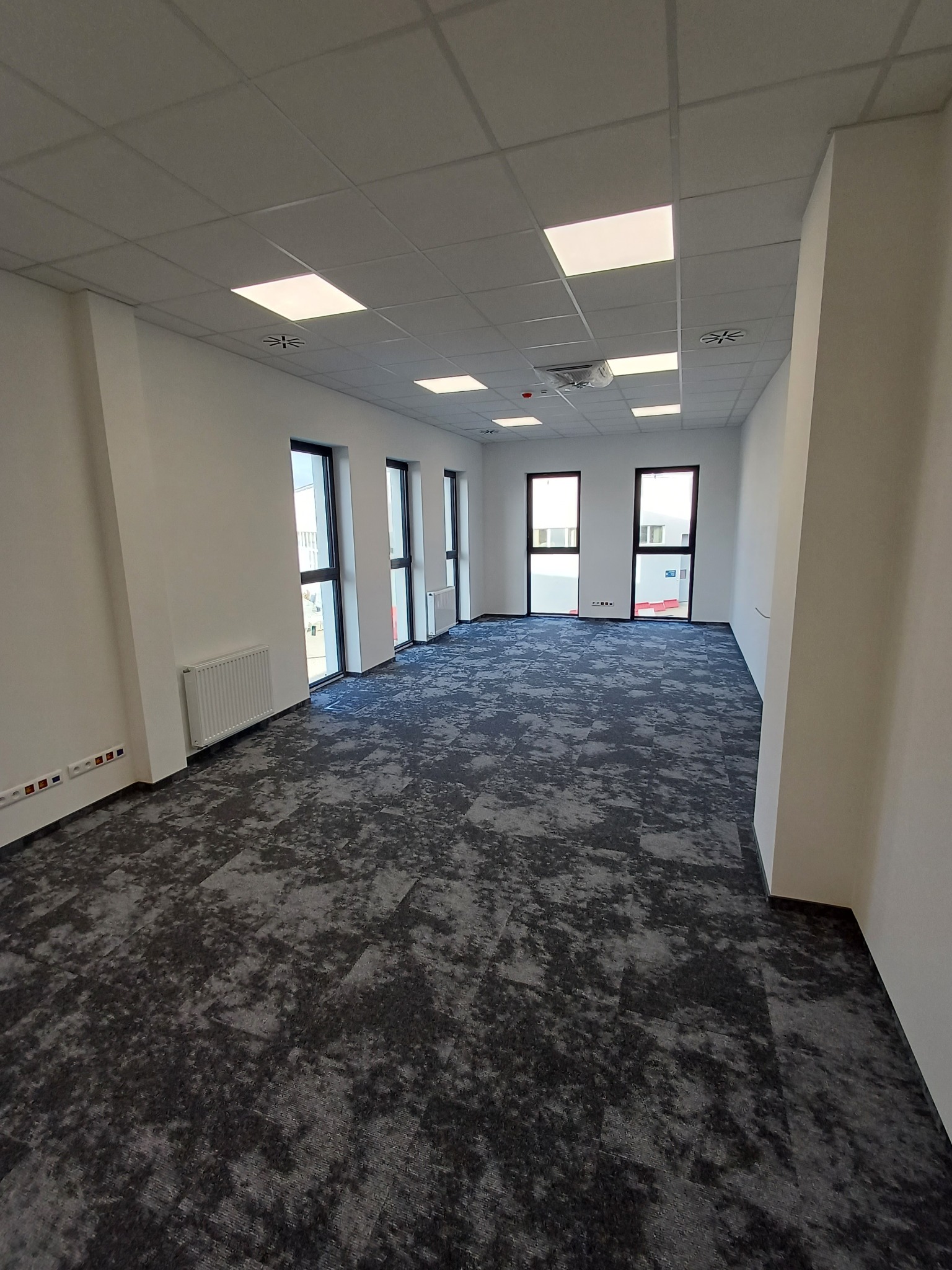Nowoczesne biuro po remoncie: szara wykładzina dywanowa, białe ściany, okna z czarnymi ramami, grzejniki i sufit kasetonowy z oświetleniem. Widok na puste, odświeżone wnętrze.