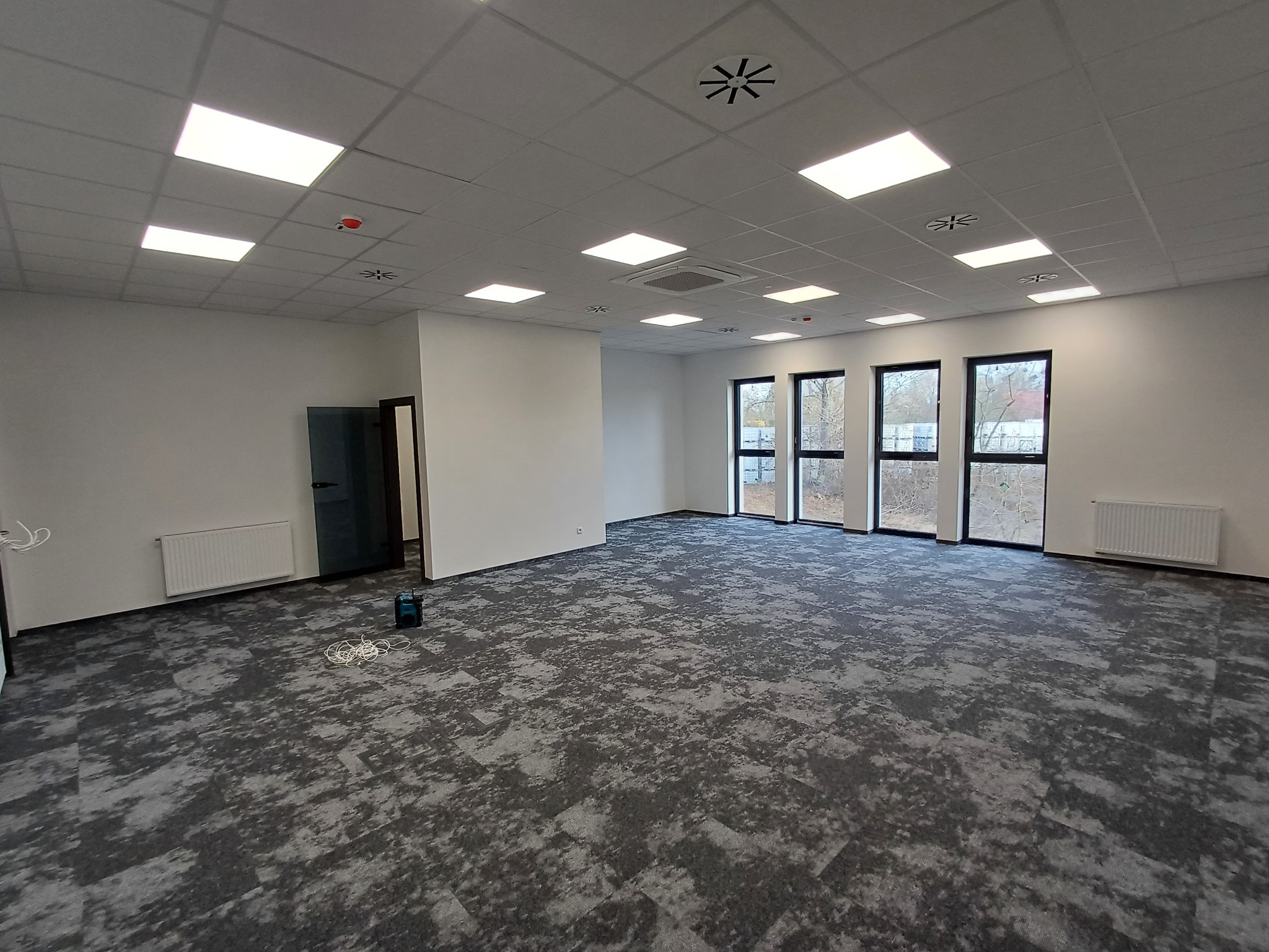 Wykończone wnętrze biurowe z szarą wykładziną dywanową w plamy, białymi ścianami i sufitami podwieszanymi z oświetleniem. Widoczne okna i grzejniki.