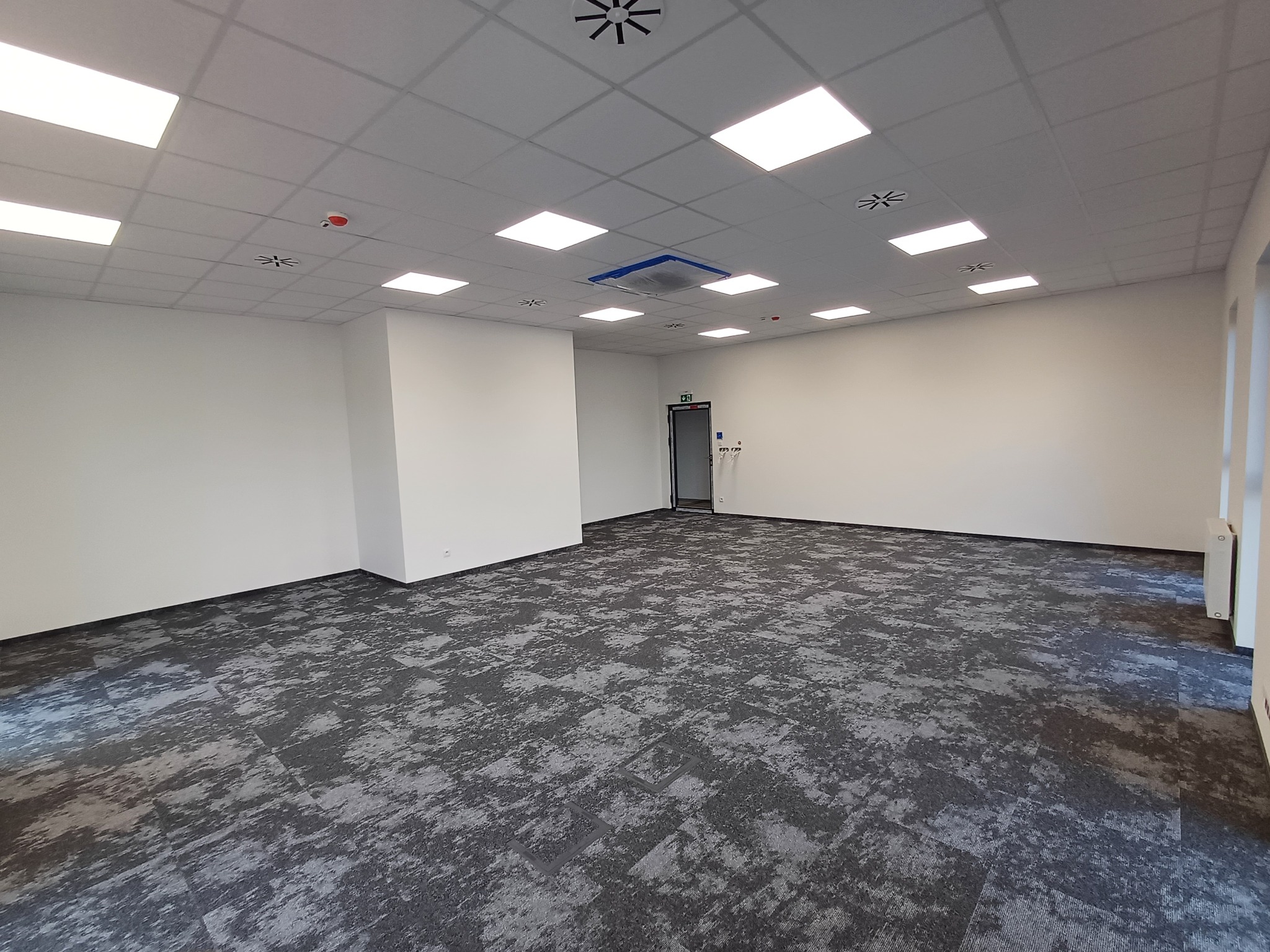 Wykończone wnętrze biurowe z szarą wykładziną dywanową w kwadraty i białymi ścianami. Sufit podwieszany z kwadratowymi lampami. Widok na całe pomieszczenie.