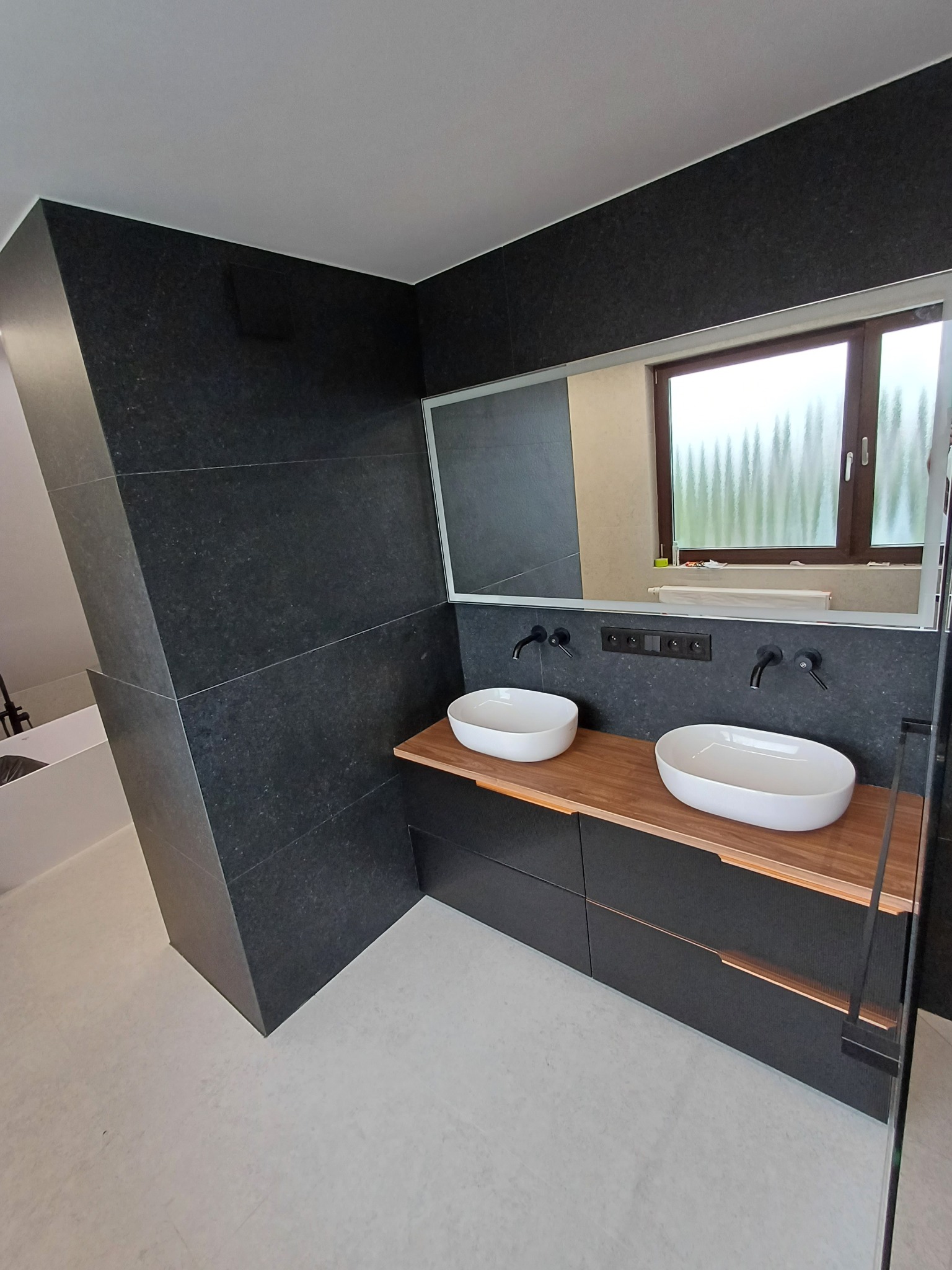 Nowoczesna łazienka z czarnymi płytkami, drewnianym blatem pod umywalki i dużym lustrem z podświetleniem. Minimalistyczny design, widok na okno z matową szybą.