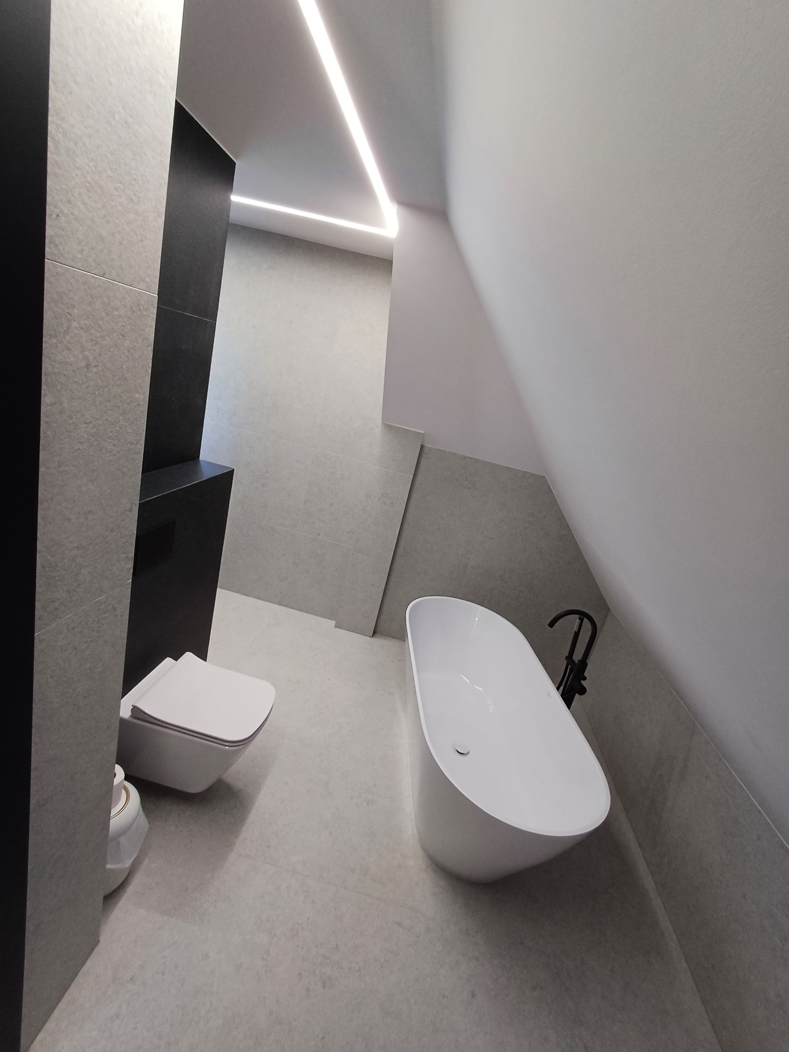 Nowoczesna łazienka w szarościach z wolnostojącą wanną i podświetleniem LED. Minimalistyczny design, płytki imitujące kamień, czarna armatura i wisząca toaleta.