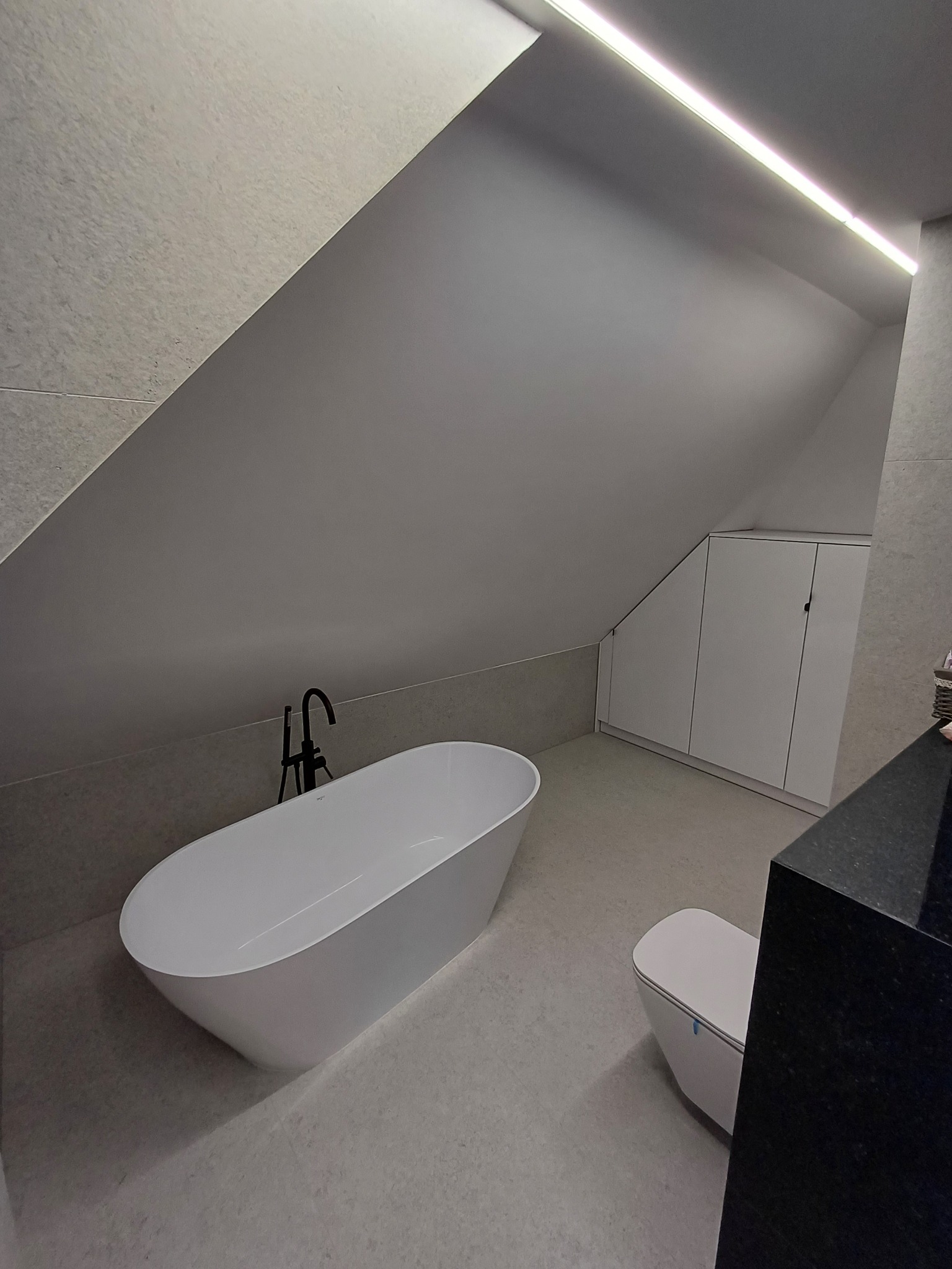 Nowoczesna łazienka na poddaszu z wolnostojącą wanną, toaletą i czarnym blatem. Białe szafki w skosie, szare płytki i oświetlenie LED tworzą minimalistyczny design.