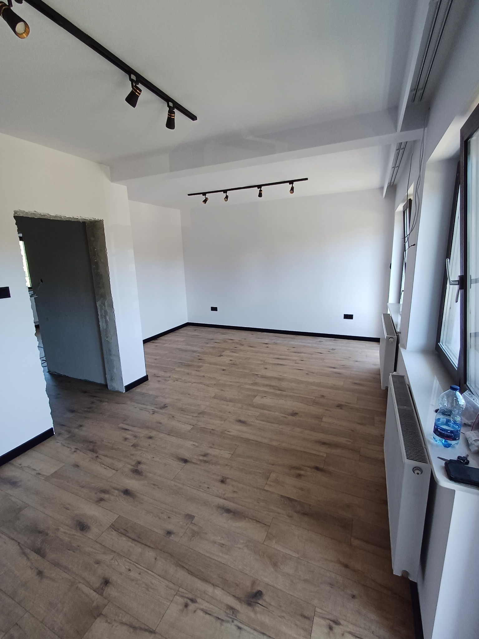 Jasne panele podłogowe w pustym, odświeżonym pokoju z białymi ścianami, czarnymi listwami i nowoczesnym oświetleniem szynowym. Widoczne okna i grzejniki.