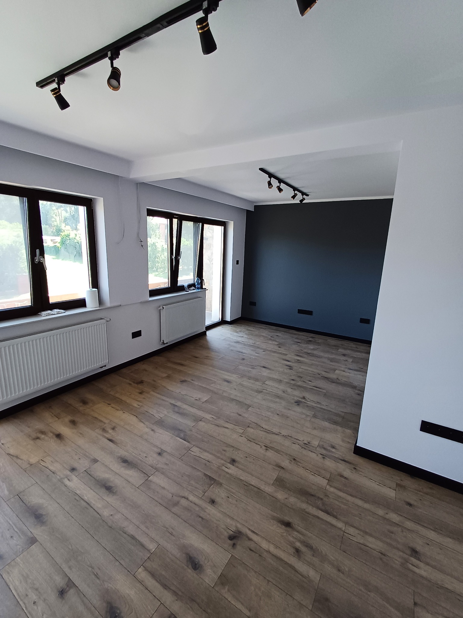 Jasne wnętrze po remoncie: panele podłogowe, szara ściana z czarnym akcentem, okna z widokiem na zieleń, nowoczesne oświetlenie szynowe. Minimalistyczny design.