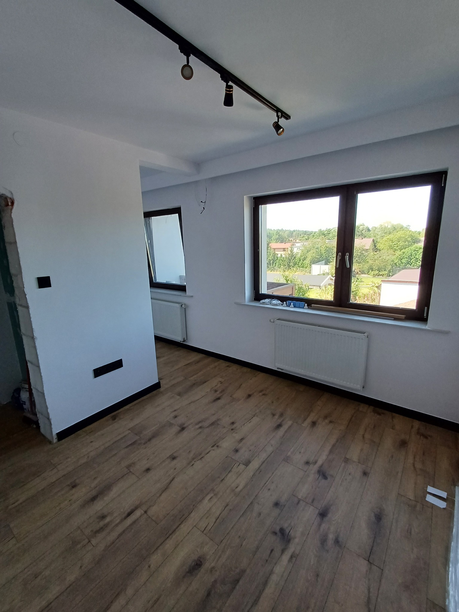 Jasne wnętrze po remoncie: panele podłogowe, białe ściany z czarnymi akcentami, okna z widokiem na zieleń. Częściowo wykończone przejście do innego pomieszczenia.