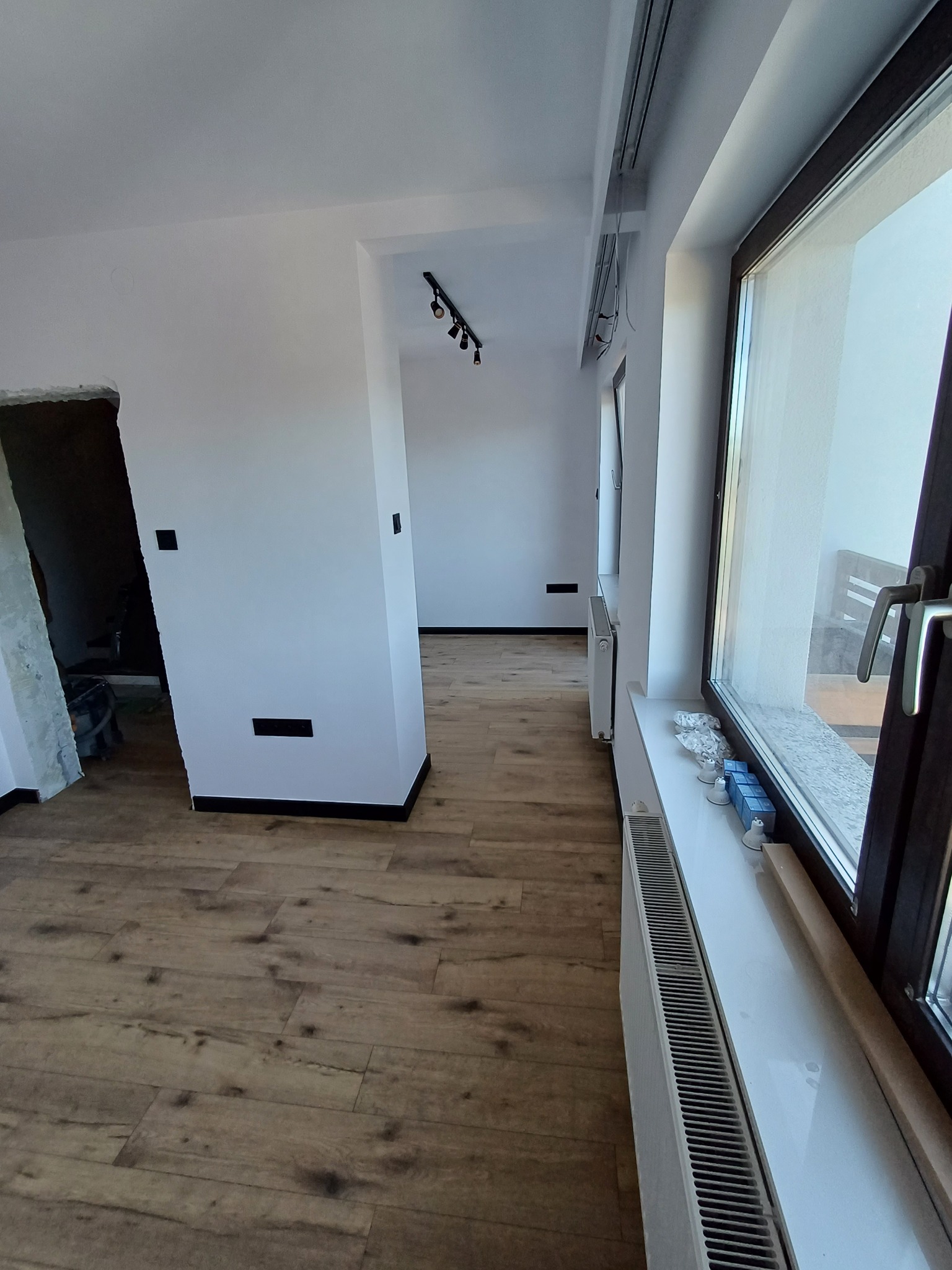 Jasne wnętrze po remoncie: drewnopodobne panele, białe ściany z czarnymi gniazdkami, grzejniki pod oknami. Widoczne ślady po pracach wykończeniowych.