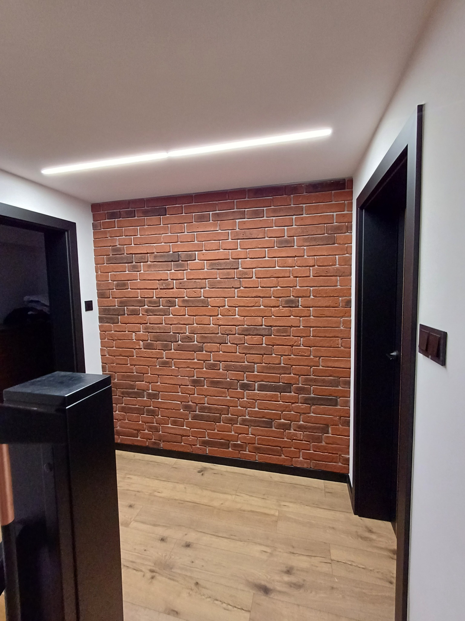 Korytarz z dekoracyjną ścianą z cegły, oświetleniem LED i panelami podłogowymi. Czarne futryny drzwi i włączniki. Widok na fragment mebla.