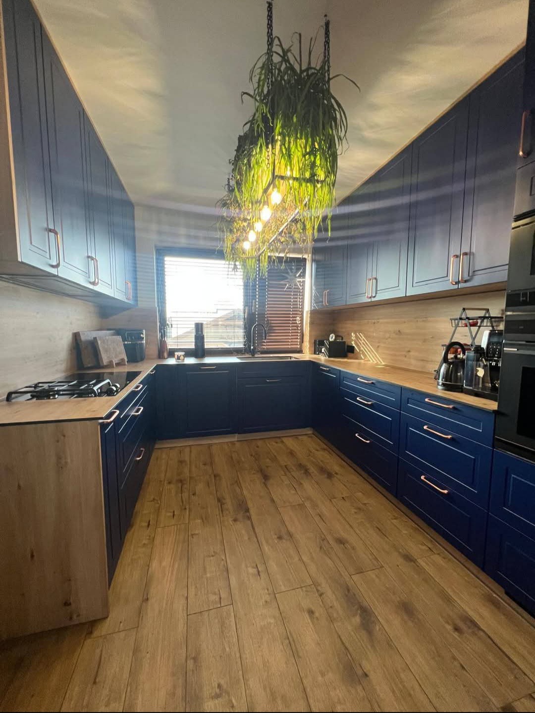 Ukończona kuchnia w odcieniach granatu i drewna, z wiszącą roślinną lampą. Widoczne szafki, blaty i drewniana podłoga. Nowoczesny design z elementami naturalnymi.