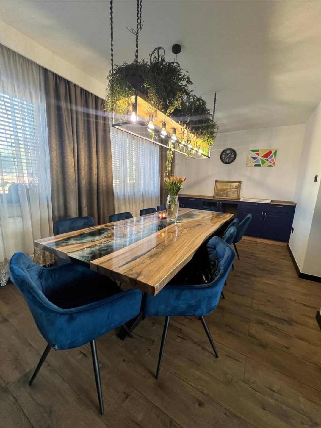 Stylowa jadalnia z drewnianym stołem z żywicą, oświetlonym wiszącą lampą z roślinami. W tle zasłony, komoda i parkiet. Nowoczesny design.