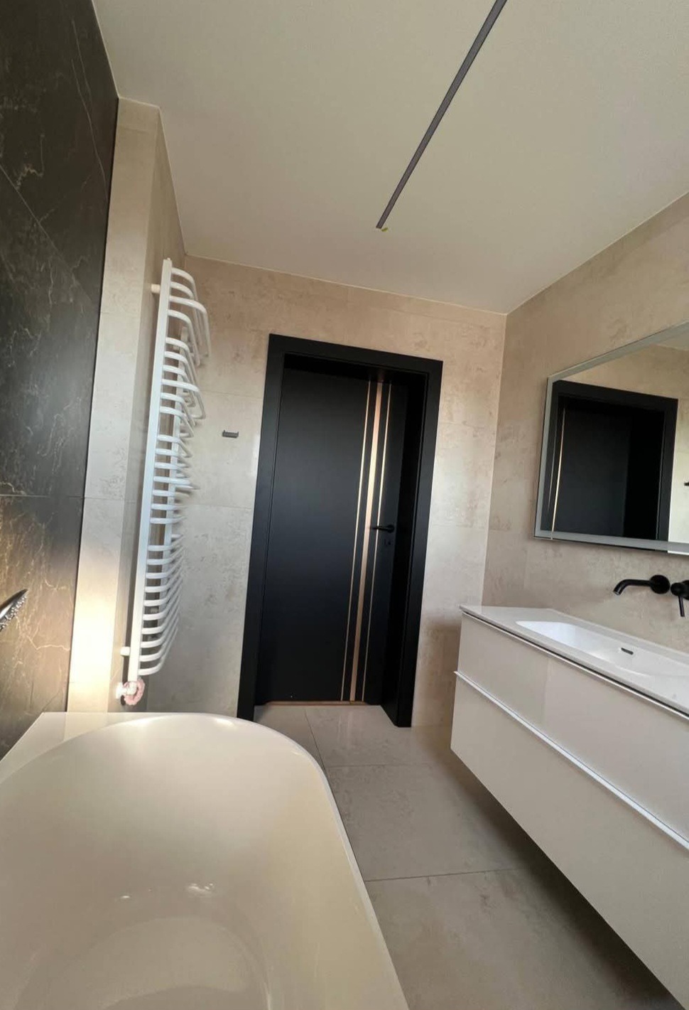 Nowoczesna łazienka z czarnymi drzwiami z złotymi paskami, białą wanną i grzejnikiem drabinkowym. Beżowe płytki i minimalistyczna umywalka z lustrem.