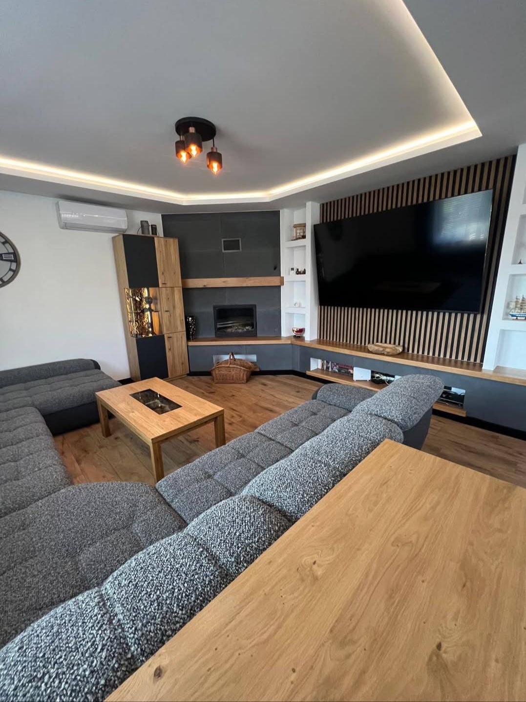 Nowoczesny salon z szarą sofą modułową, drewnianą zabudową TV i kominkiem. Oświetlenie LED podkreśla geometrię wnętrza. Minimalistyczny design.