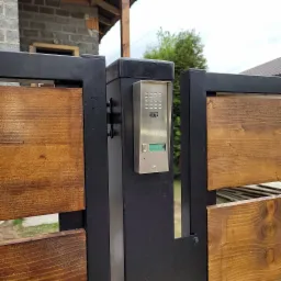 Metalowa brama z drewnianymi panelami i zamontowanym srebrnym wideofonem Urmet Miwi z zielonym wyświetlaczem.
