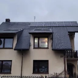 Dwukondygnacyjny dom z zamontowanymi panelami słonecznymi na dachu z blachy trapezowej, antena satelitarna, numer domu 23, pochmurne niebo.