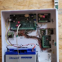 Otwarta skrzynka z elektroniką systemu alarmowego, widoczne płyty główne, okablowanie, akumulator Ultracell i transformator Pulsar AWT500, zamontowana na płycie OSB.
