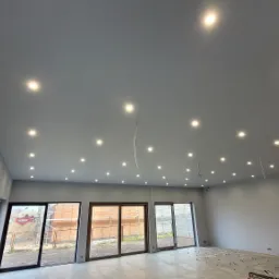 Szare wnętrze z licznymi punktowymi lampami LED w suficie, widoczne niepodłączone przewody i okna z widokiem na budowę.