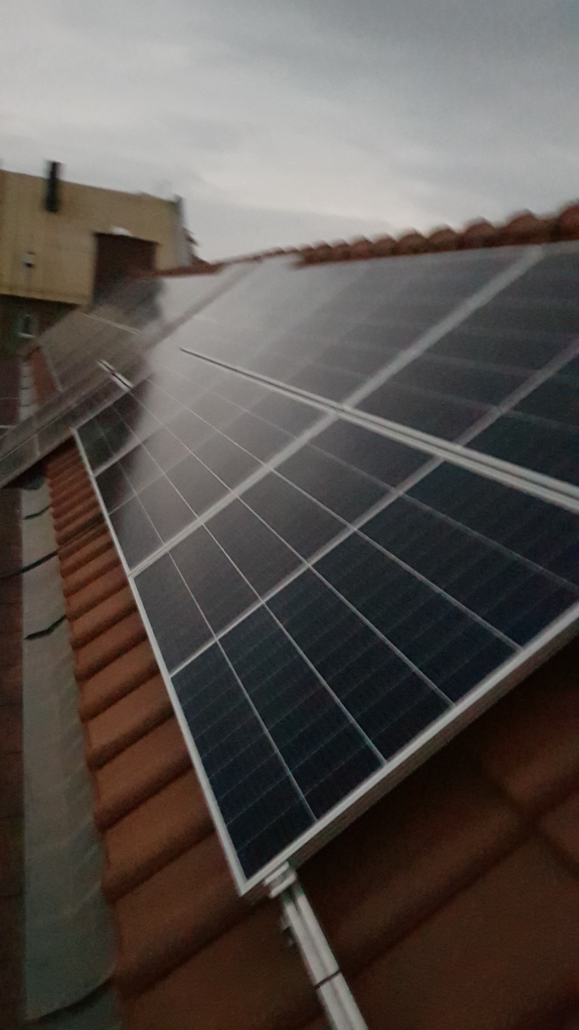 Panel słoneczny zamontowany na brązowym dachu z dachówki ceramicznej, widok z dołu pod kątem, pochmurne niebo w tle.