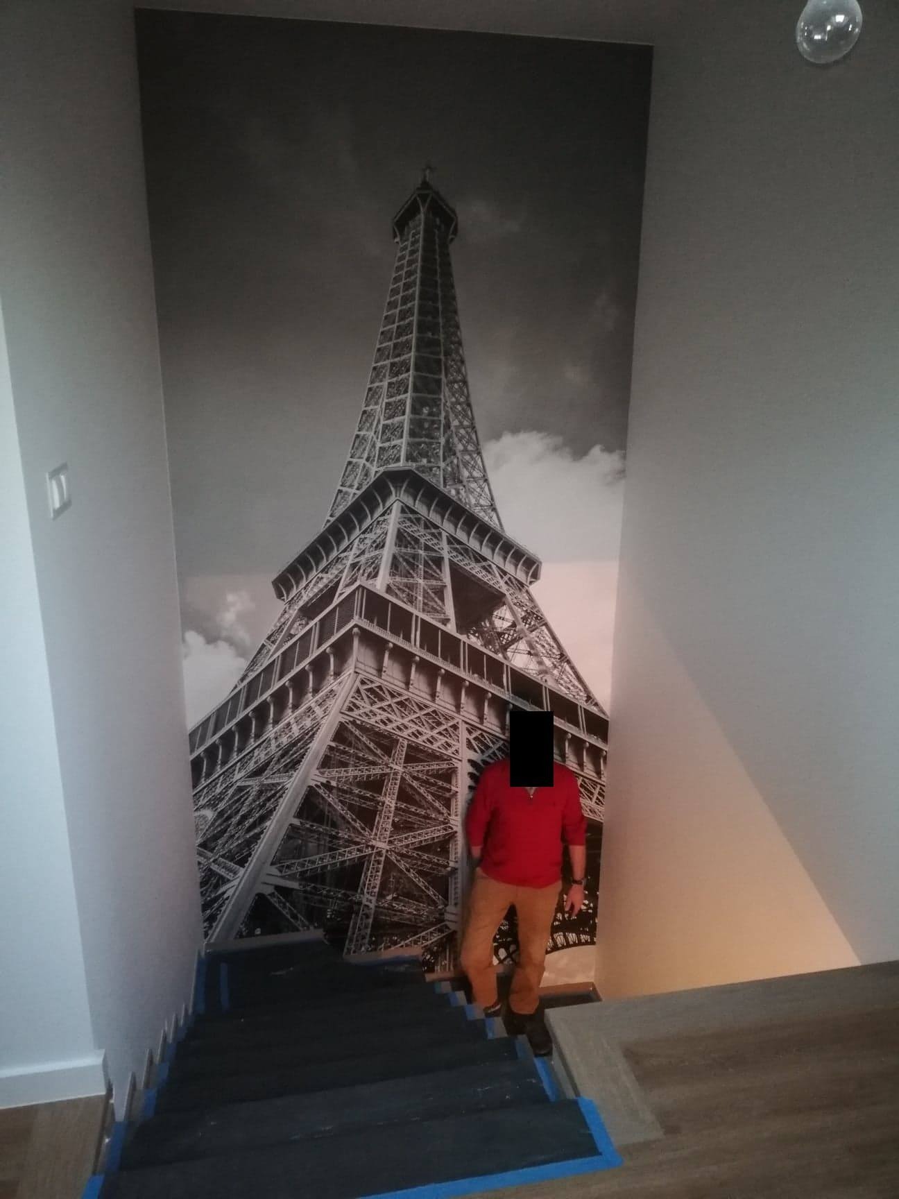 Czarno-biała fototapeta z wieżą Eiffla na ścianie klatki schodowej, osoba w czerwonej bluzie schodzi po schodach.