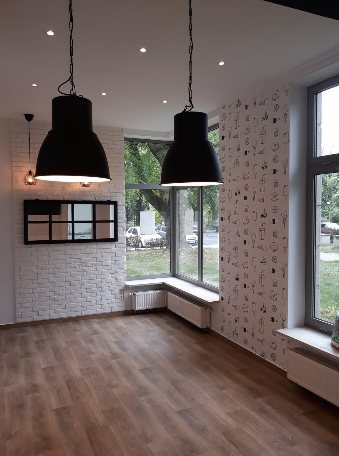 Jasne wnętrze lokalu z drewnianą podłogą, białymi ceglanymi ścianami, dużymi oknami, grzejnikami pod oknami i dwoma dużymi czarnymi lampami wiszącymi, jedna ściana ozdobiona wzorem lodów i napojów.
