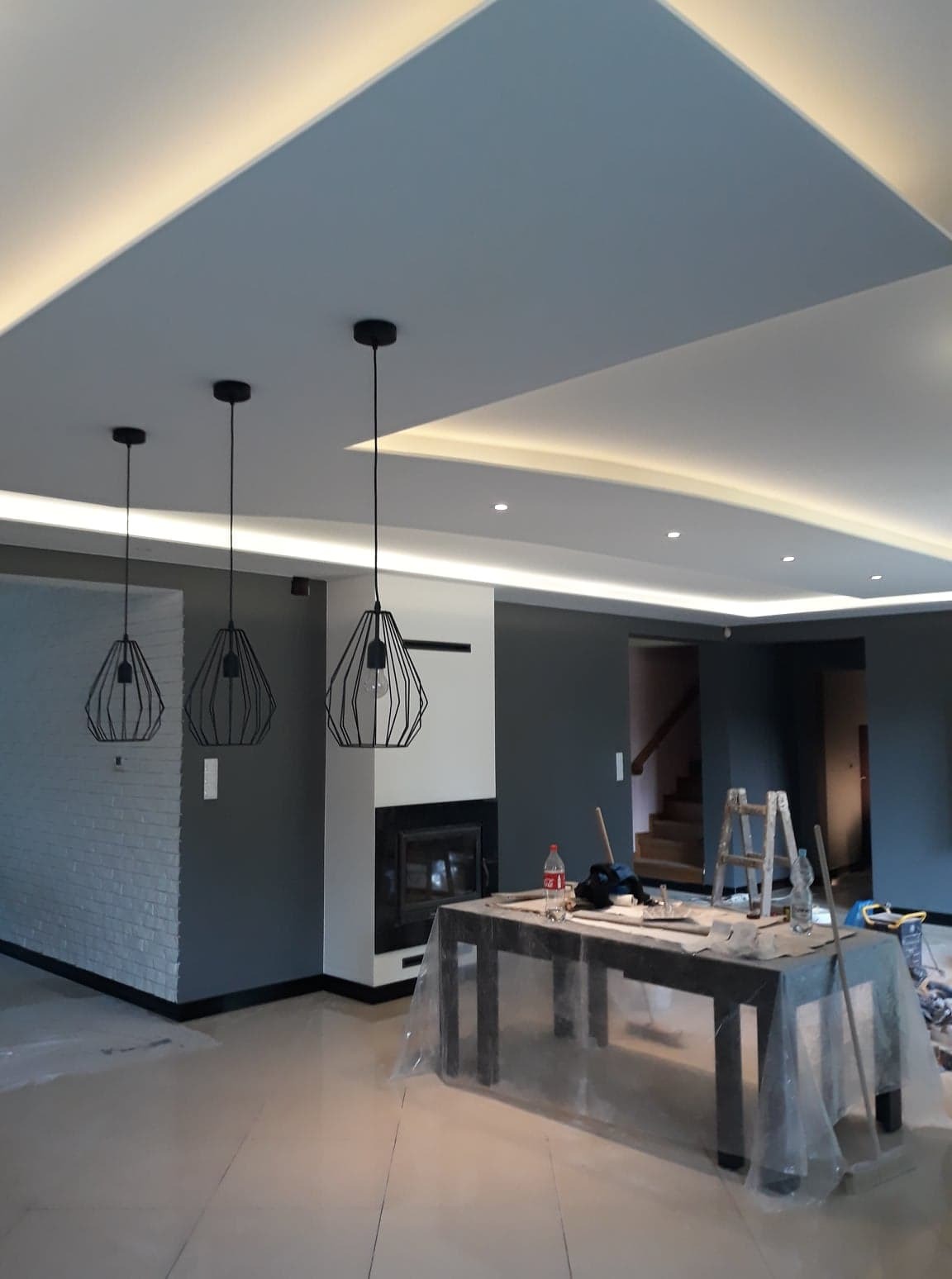 Salon w trakcie wykończenia: pomalowane ściany na szaro, sufit z podświetleniem LED, wiszące lampy druciane, kominek i zabezpieczone folią meble.