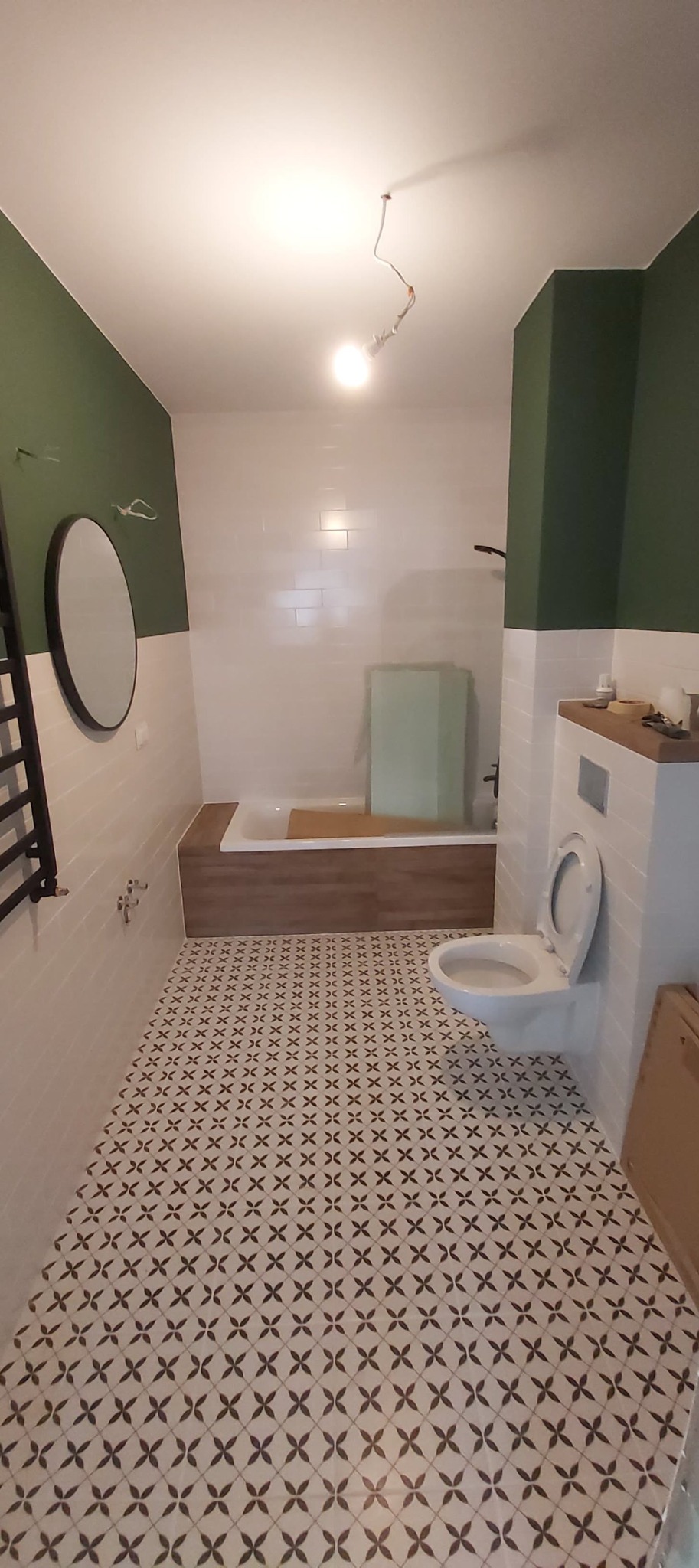 Wnętrze łazienki po remoncie z wzorzystą podłogą, białą armaturą, obudowaną wanną w kolorze drewna i ścianami w tonacji bieli i butelkowej zieleni, z niedokończonym oświetleniem sufitowym.