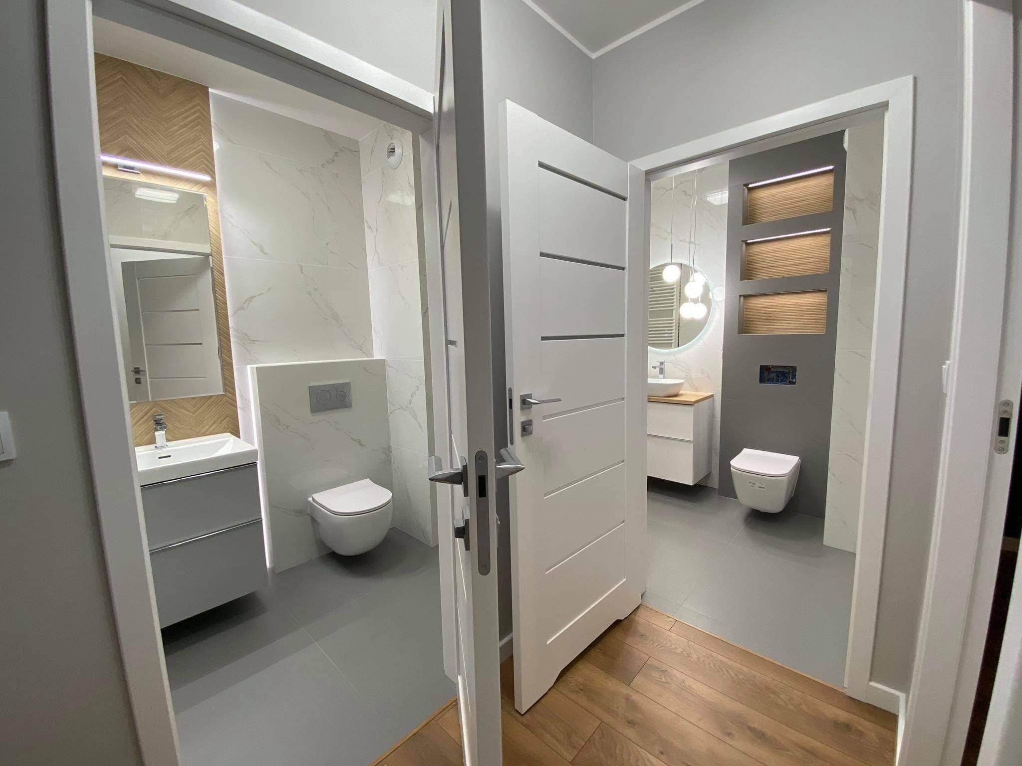 Widok na dwie łazienki po remoncie, z drewnianą podłogą na pierwszym planie i otwartymi białymi drzwiami, ukazującymi szare płytki, białą armaturę i nowoczesne oświetlenie.