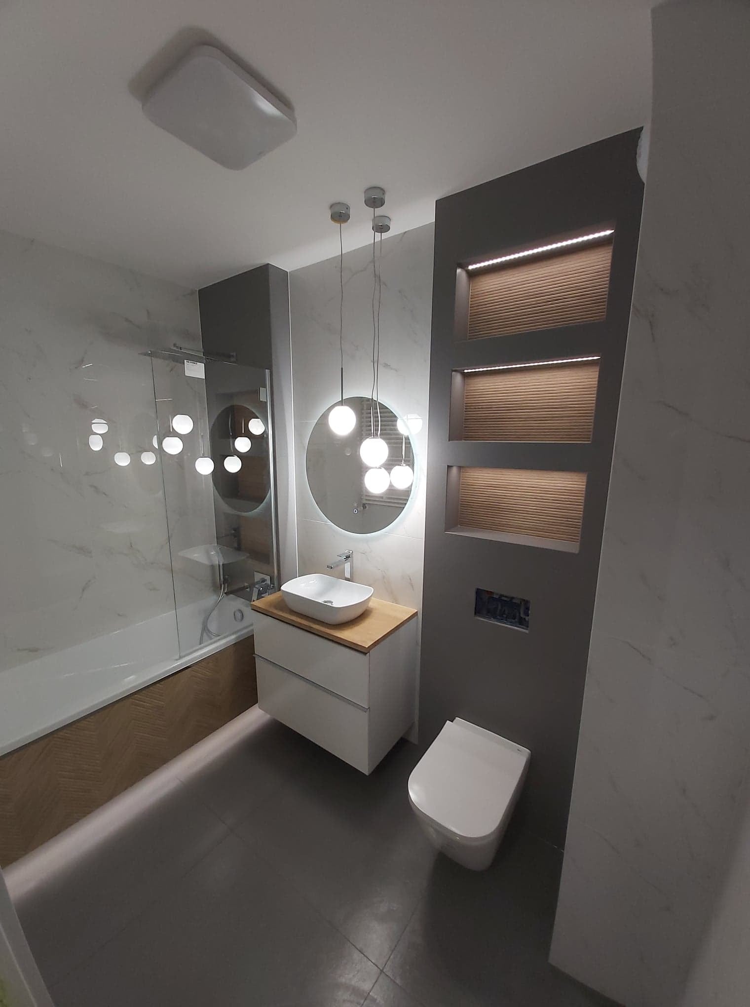 Nowoczesna łazienka z marmurowymi płytkami, okrągłym lustrem z podświetleniem LED, wiszącą szafką z umywalką nablatową, podwieszaną toaletą i dekoracyjną ścianą z podświetlanymi wnękami.