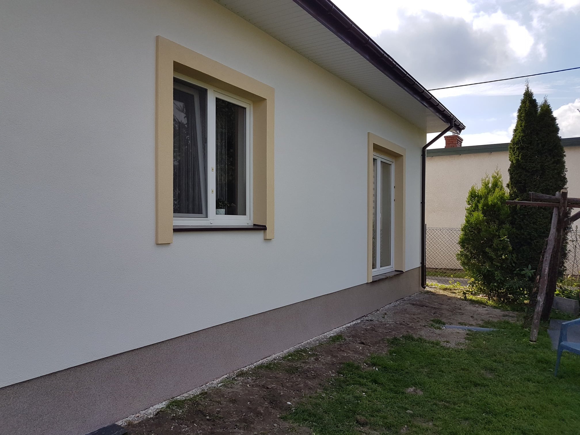 Elewacja domu jednorodzinnego po remoncie, widoczne ocieplenie ścian i odmalowane okna z kremowymi ramami, jasna elewacja i ciemniejsza podmurówka.