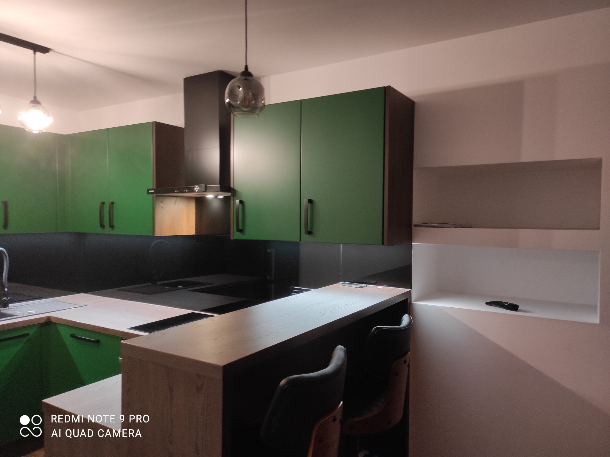 Nowoczesna kuchnia z zielonymi szafkami, czarnym panelem nad blatem, drewnianym barkiem i dwoma hokerami. Widoczna wisząca lampa z szklanym kloszem i wnęka w ścianie z pilotem.