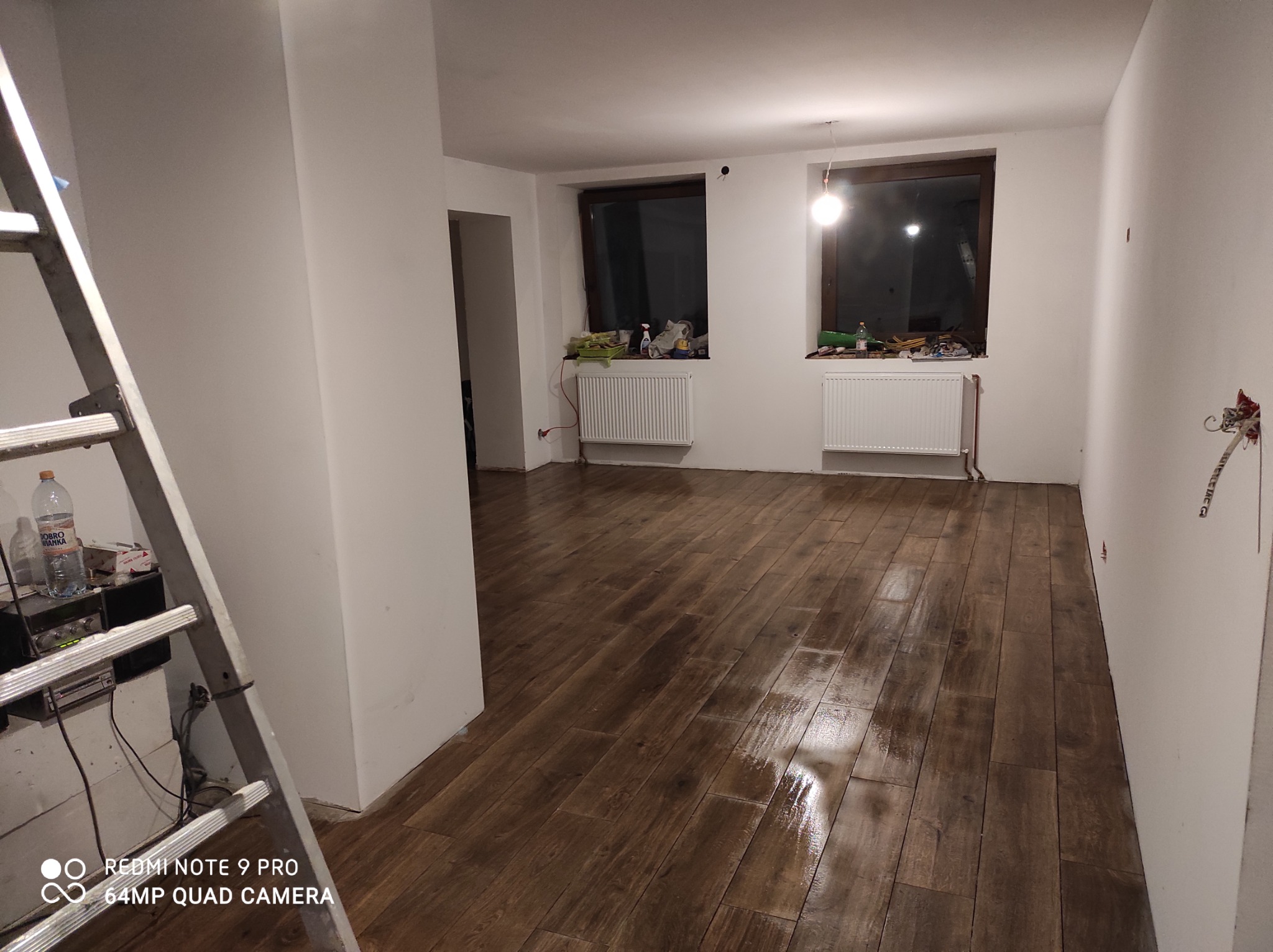 Wykończone wnętrze z brązową podłogą imitującą drewno, białe ściany i grzejniki pod oknami, widoczna drabina malarska z butelką wody mineralnej.