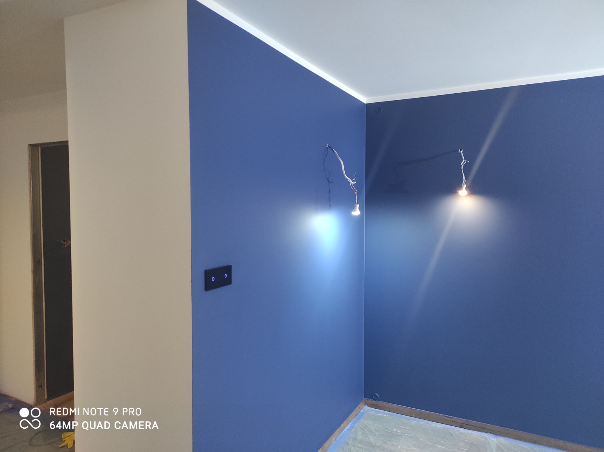 Świeżo pomalowane na niebiesko ściany w rogu pokoju z widocznymi przewodami elektrycznymi pod lampy i czarnym włącznikiem światła, zabezpieczona folią podłoga.