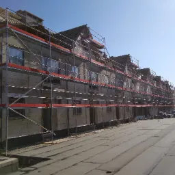 Budynek wielorodzinny w trakcie prac budowlanych, zabezpieczony rusztowaniami z czerwonymi elementami, widok na elewację i dach w surowym stanie, błękitne niebo w tle.