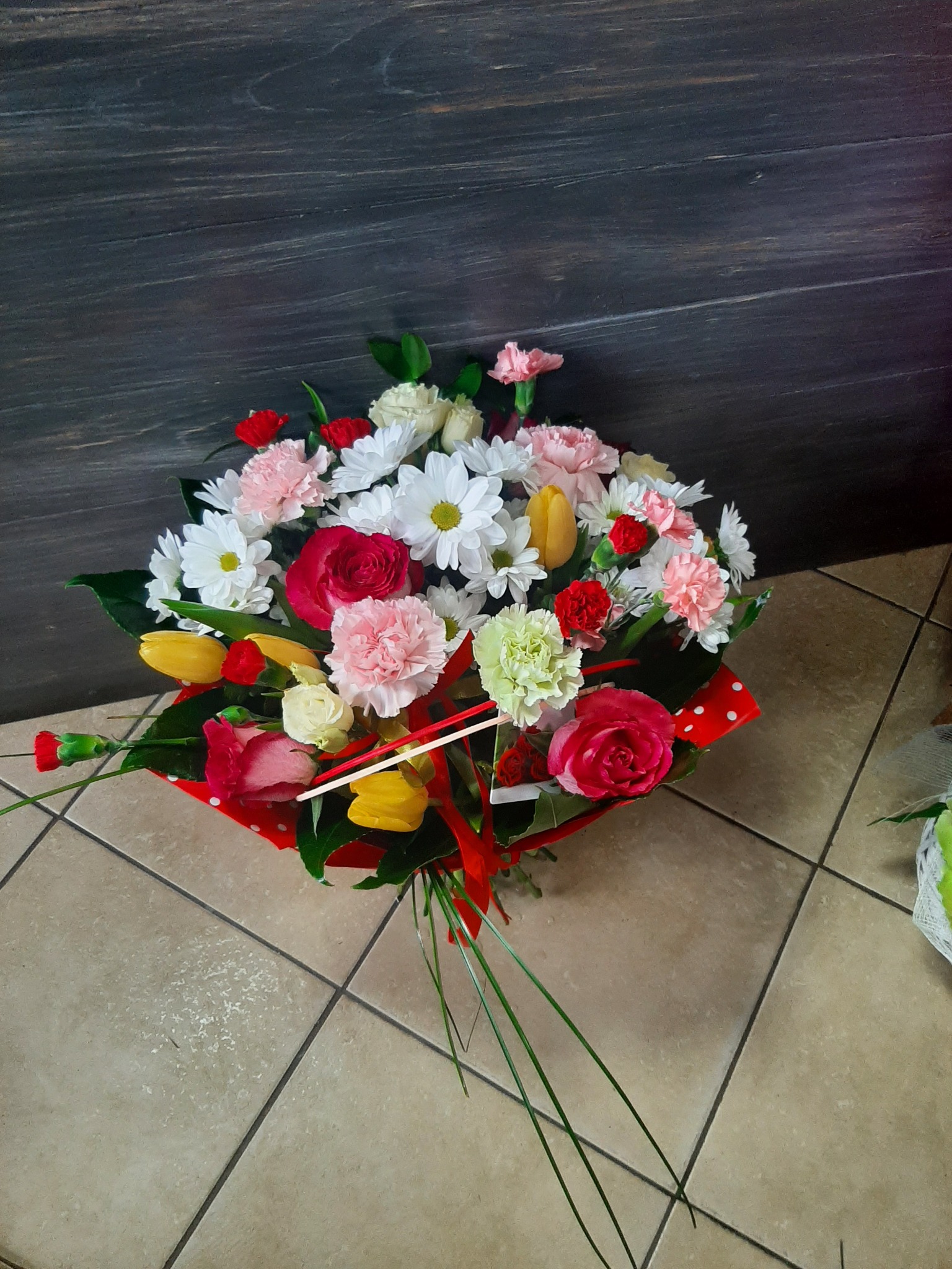 Kompozycja kwiatowa w czerwonym papierze w białe kropki, zawierająca róże, goździki, tulipany i margerytki, ułożona na kafelkach podłogowych na tle ciemnej ściany.