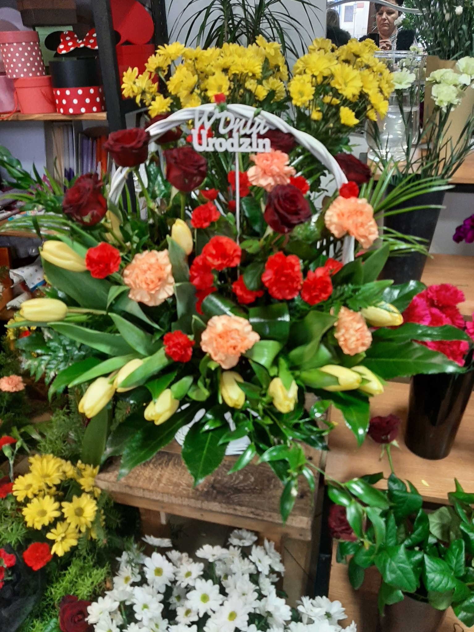 Kosz z mieszanką kwiatów: czerwone róże, czerwone i pomarańczowe goździki, żółte tulipany, dekorowany napisem 'W Dniu Urodzin', umieszczony na drewnianym podeście w kwiaciarni, w tle inne kwiaty...