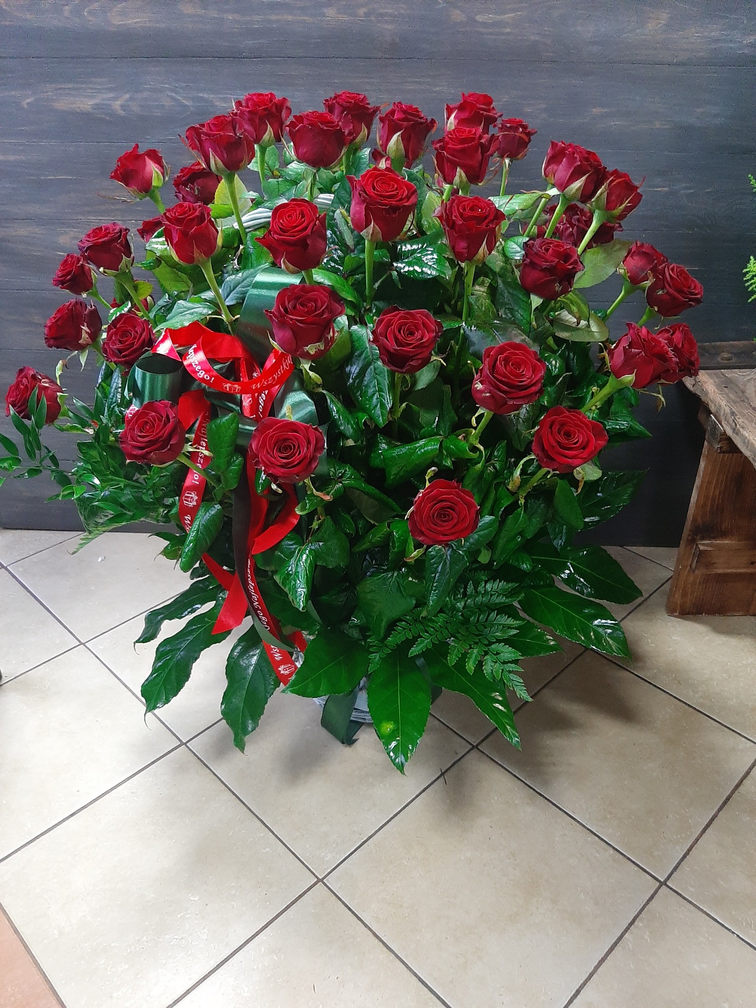 Duży, okrągły bukiet z czerwonych róż, zielonych liści i wstążek z napisem, ustawiony na kafelkowej podłodze, z drewnianym panelem i fragmentem stołka w tle.