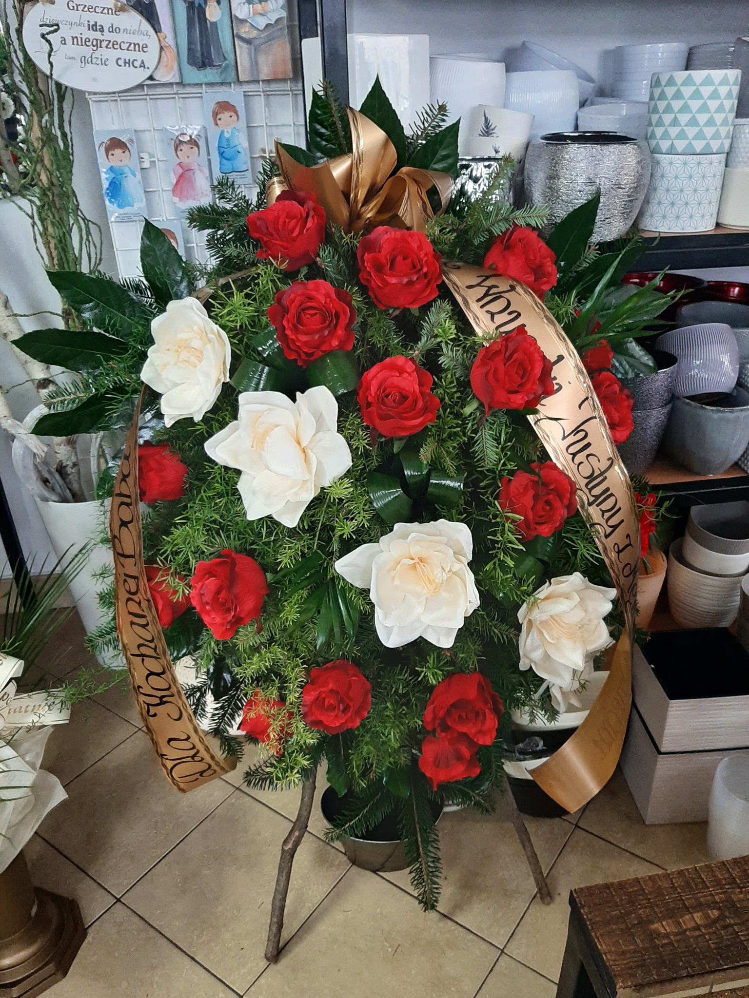Wieniec pogrzebowy z czerwonych i białych róż, zielonych gałązek i złotej wstęgi z napisem, ustawiony na drewnianym stojaku w kwiaciarni, w tle doniczki i dekoracje.