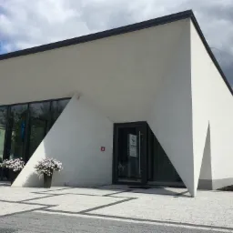 Nowoczesny budynek kliniki z charakterystycznym, trójkątnym elementem architektonicznym nad wejściem, ozdobiony kwiatami w donicach i niskimi krzewami, z szarą kostką brukową na podjeździe.