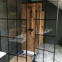 Nowoczesna kabina prysznicowa z czarnymi profilami i szklanymi ściankami, widoczne płytki podłogowe o wzorzystym deseniu oraz drewniana obudowa prysznica z deszczownicą, w tle umywalka.