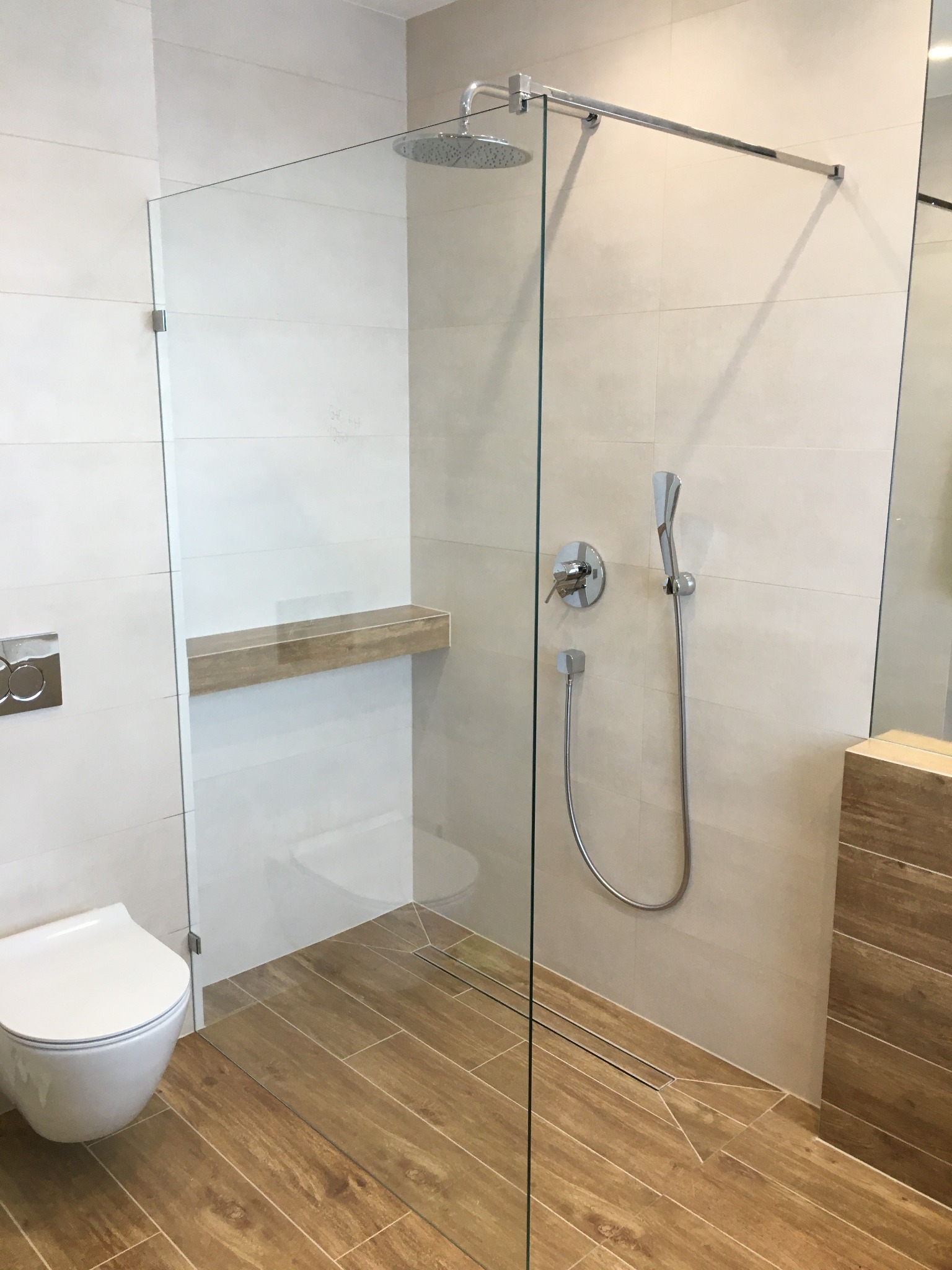 Nowoczesna kabina prysznicowa ze szklaną ścianą, deszczownicą i półką w jasnej łazience z drewnopodobnymi płytkami na podłodze.