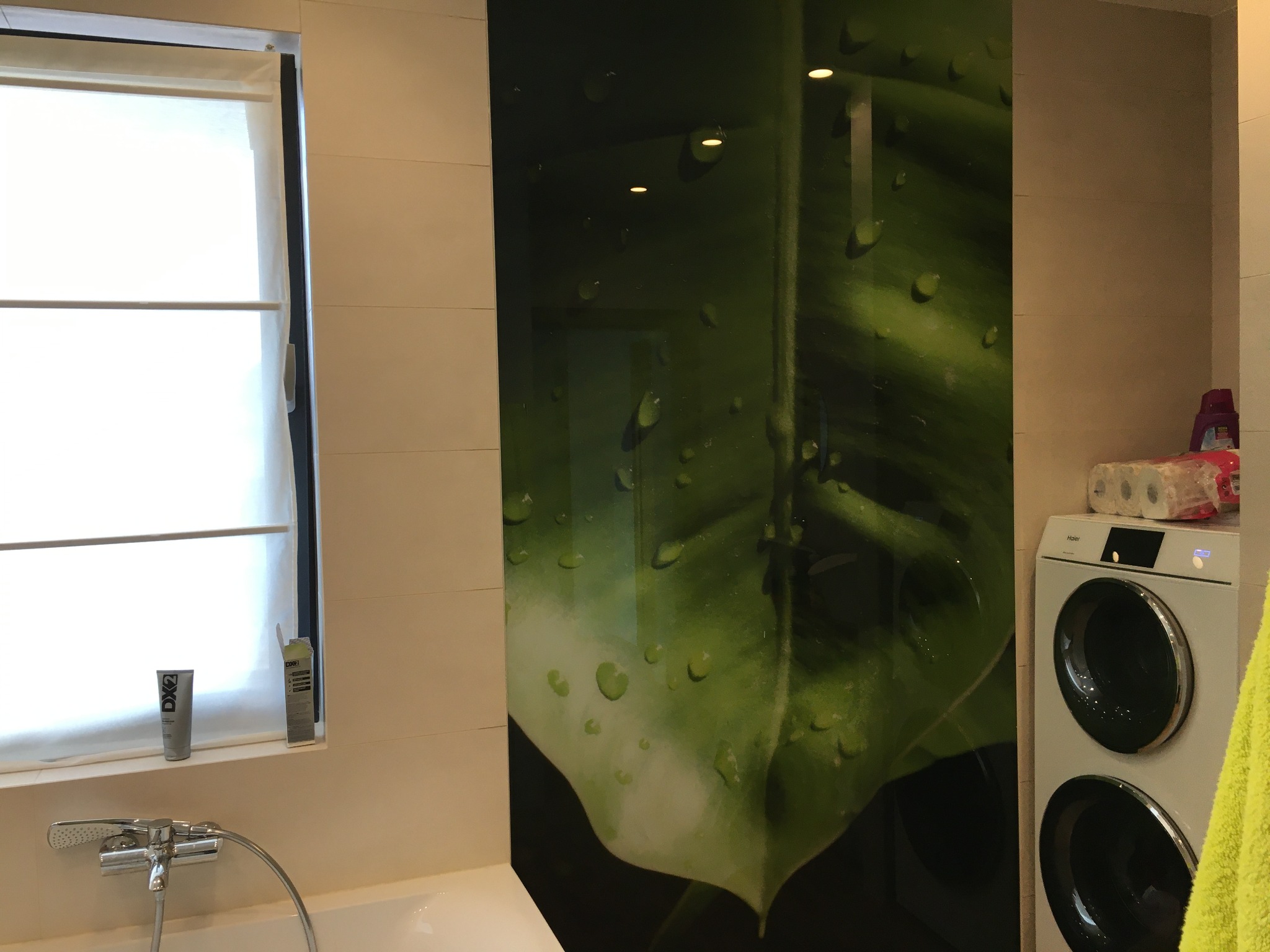 Szklany panel z motywem liścia w łazience, widoczna pralka i okno z roletą.