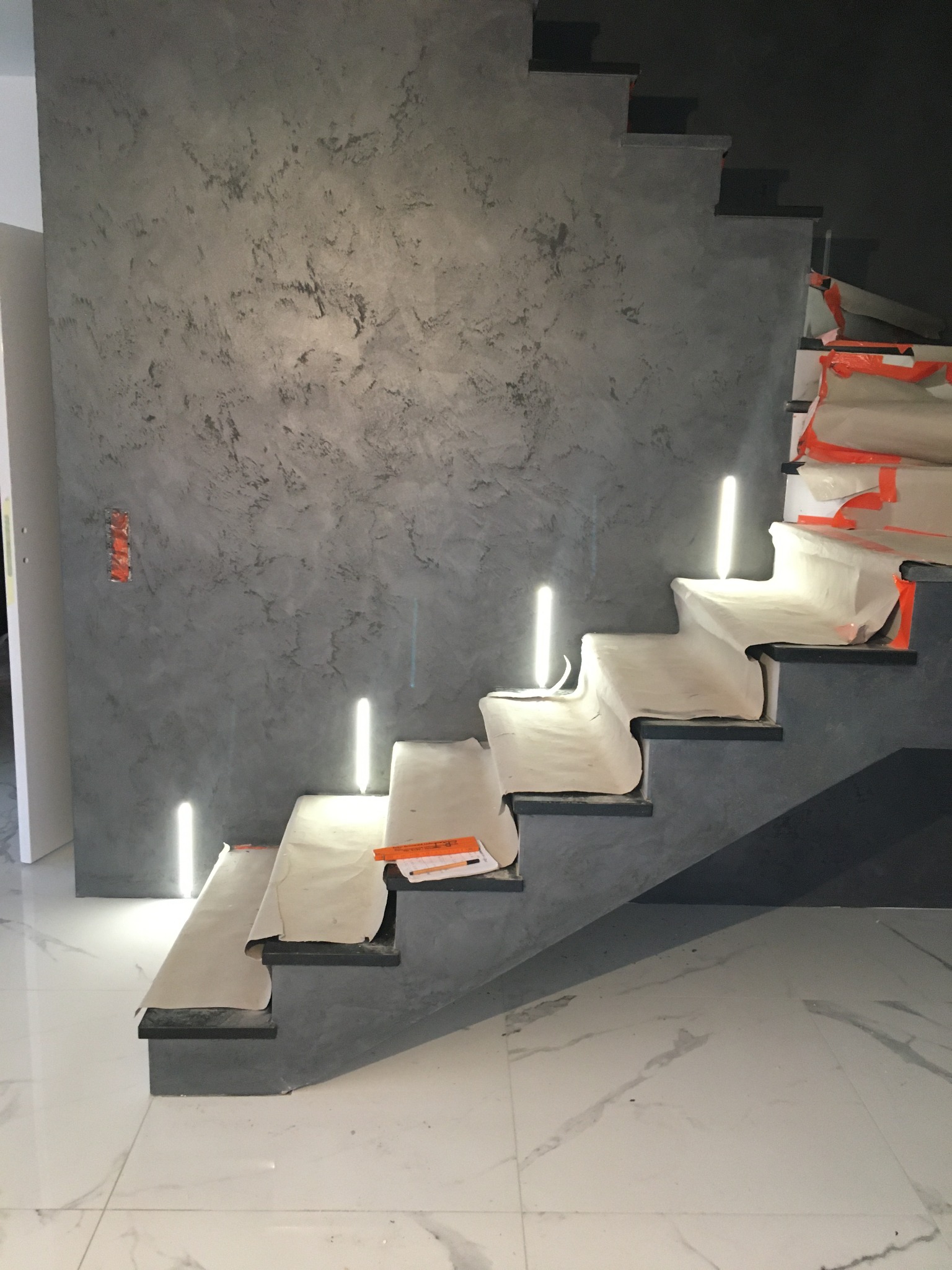 Schody betonowe z wbudowanym oświetleniem LED w trakcie zabezpieczania folią malarską przed pracami wykończeniowymi ściany o fakturze betonu architektonicznego.