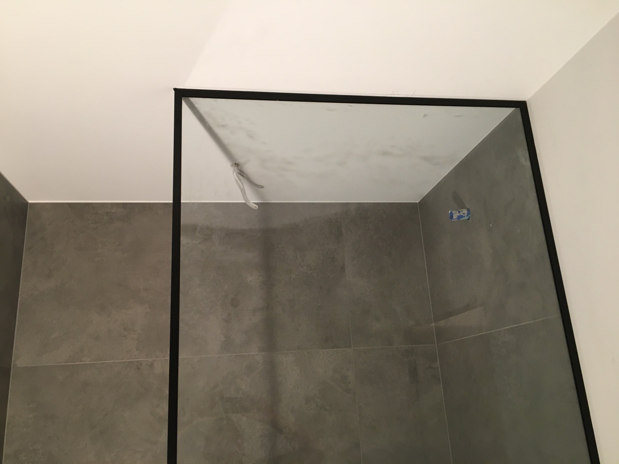 Szklana kabina prysznicowa z czarną ramą, zamontowana w rogu łazienki wyłożonej szarymi płytkami. Widoczne pozostałości po montażu na suficie.