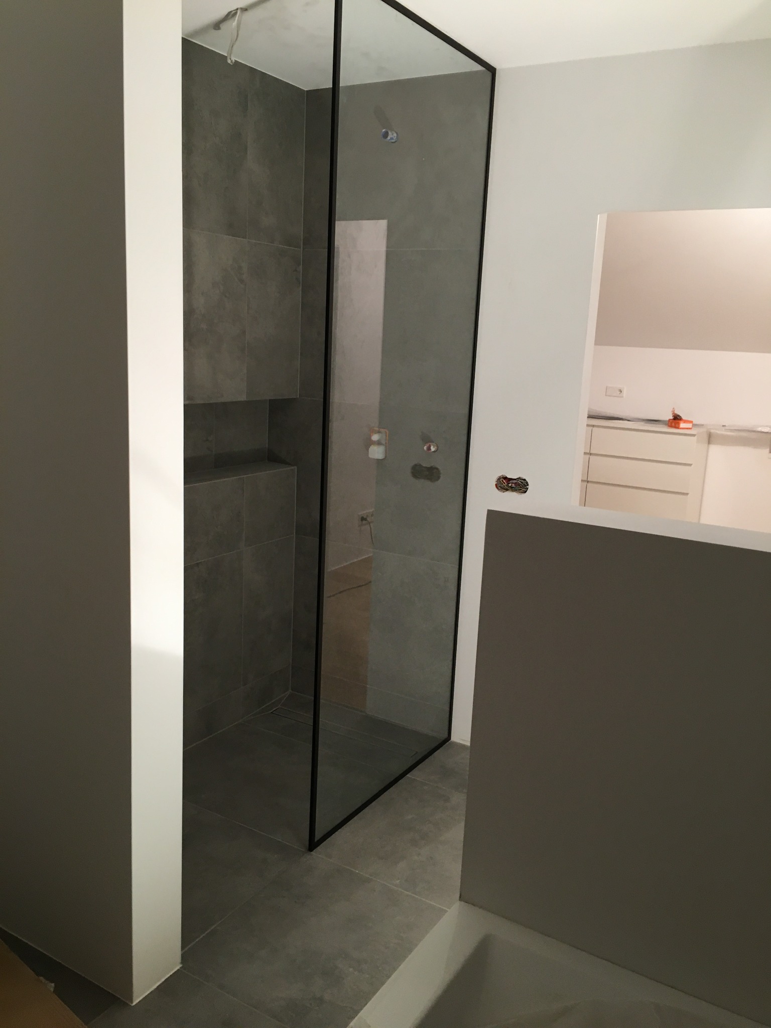 Nowoczesna kabina prysznicowa ze szklaną ścianą i czarną ramą, szare płytki wewnątrz, widoczne elementy instalacji elektrycznej na ścianie obok.