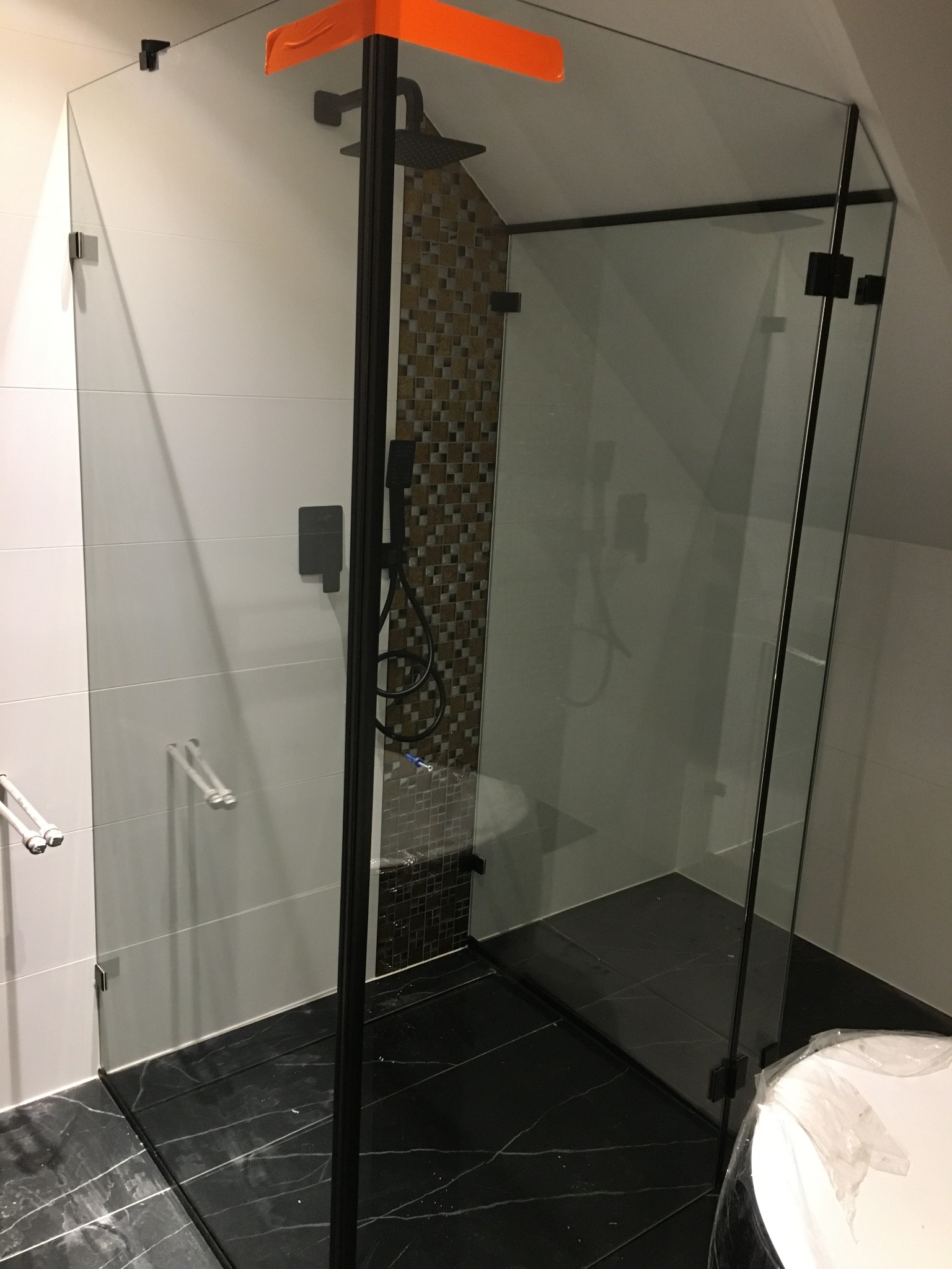 Nowoczesna kabina prysznicowa ze szklanymi ścianami i czarnymi okuciami, widoczna pomarańczowa taśma zabezpieczająca na górnej krawędzi, czarna marmurowa podłoga i częściowo widoczna wanna w folii...