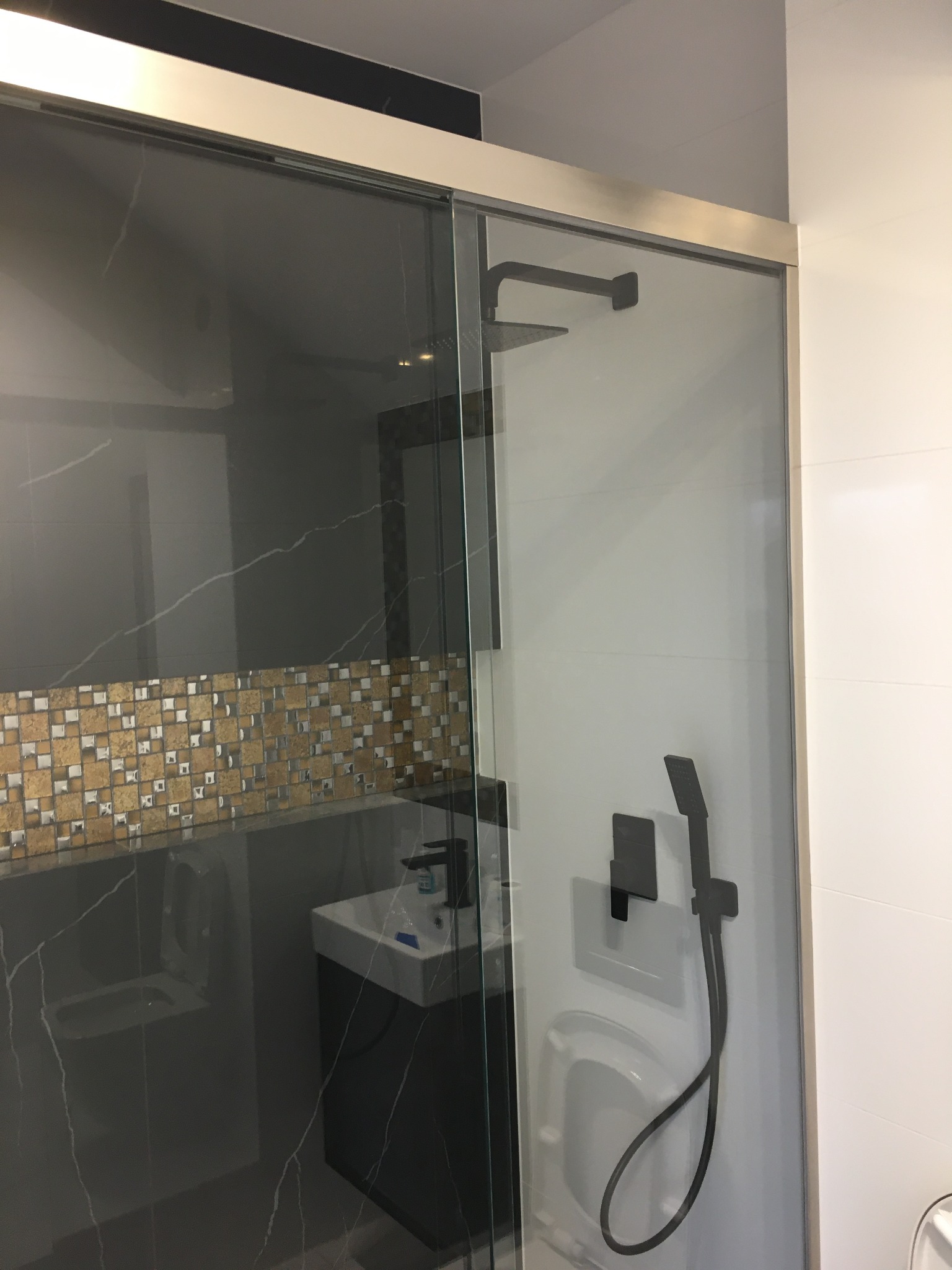 Nowoczesna kabina prysznicowa z przesuwnymi drzwiami szklanymi, czarną armaturą i mozaiką na ścianie w tle.