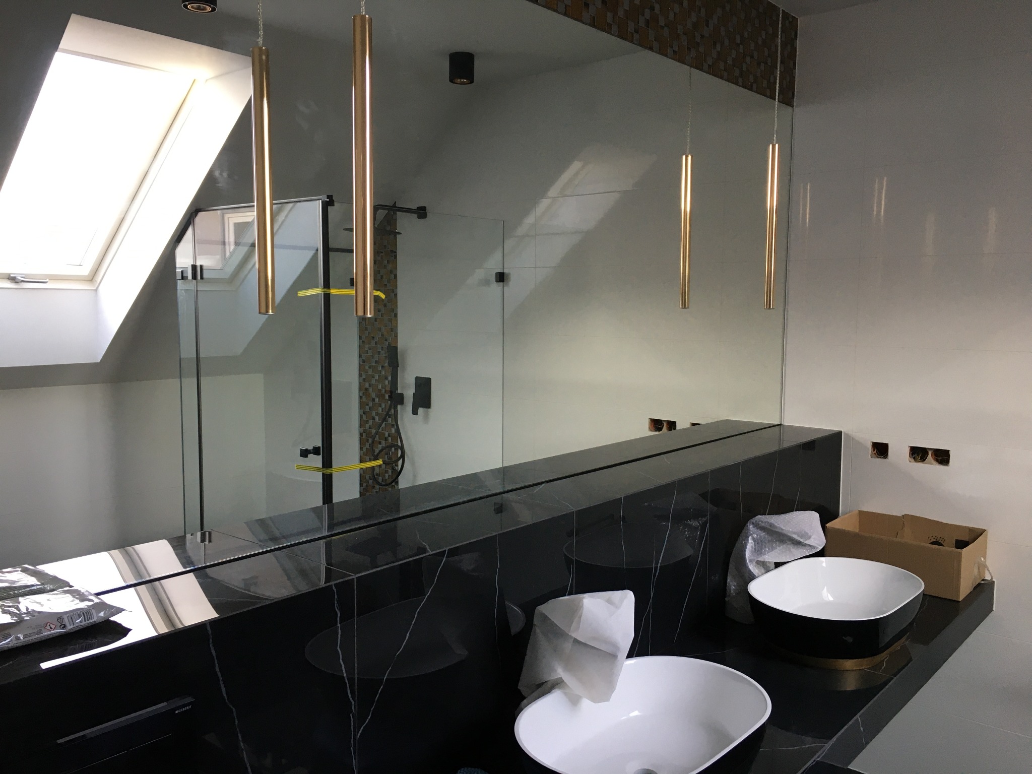 Nowoczesna łazienka w trakcie wykańczania: duże lustro z odbiciem kabiny prysznicowej, dwie białe umywalki nablatowe na czarnym blacie z marmurowym wzorem, wiszące złote lampy tuby.