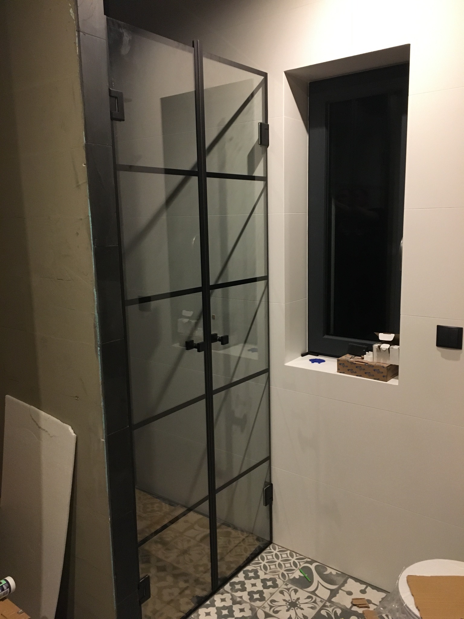 Szklana kabina prysznicowa z czarnymi profilami i ramami, zamontowana w łazience z mozaikową podłogą i białymi płytkami ściennymi, widoczne niedokończone prace wykończeniowe na ścianie obok.